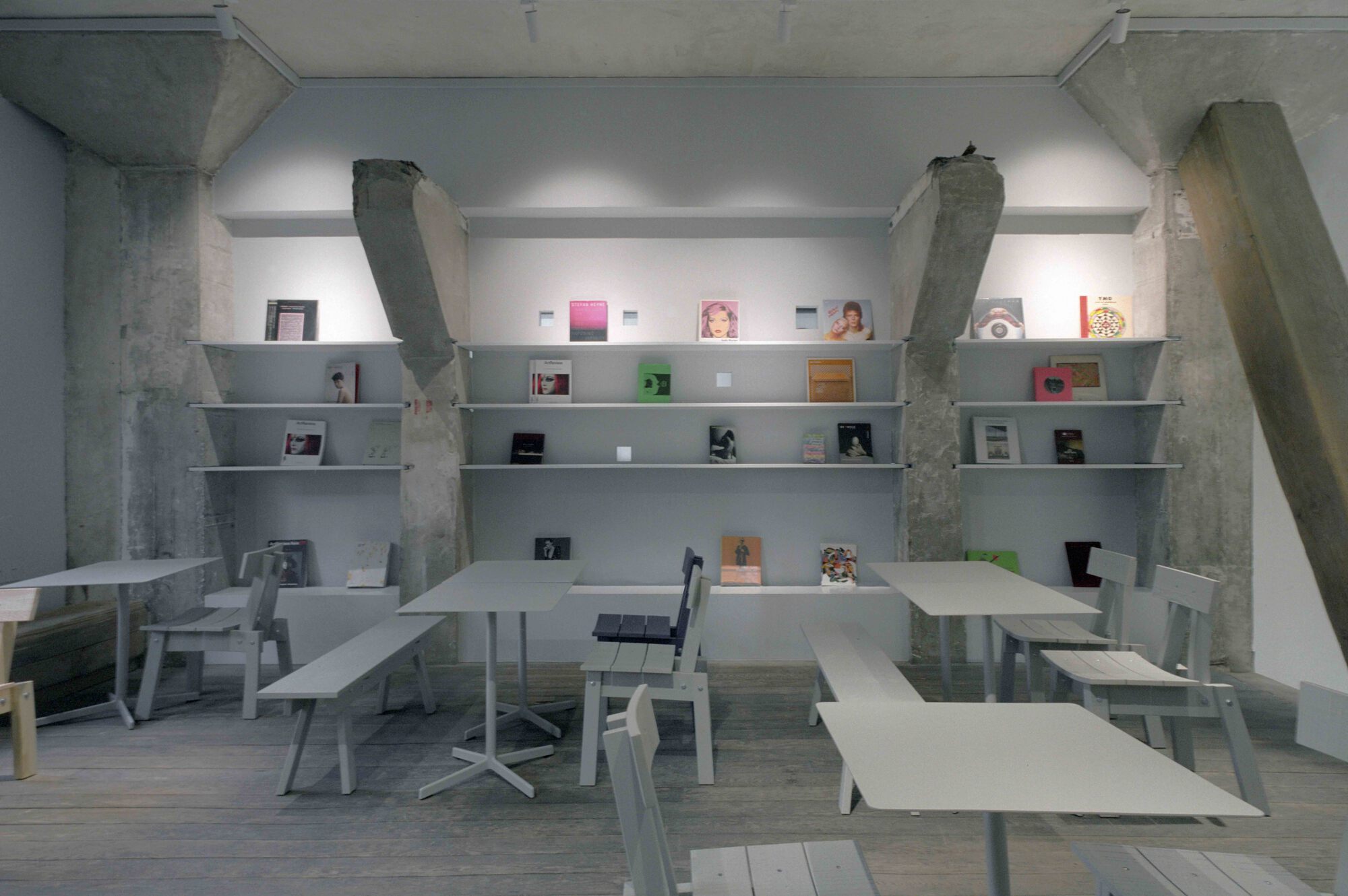 Interior workspace with white metal tables and benches beneath wall-mounted shelving and exposed concrete columns