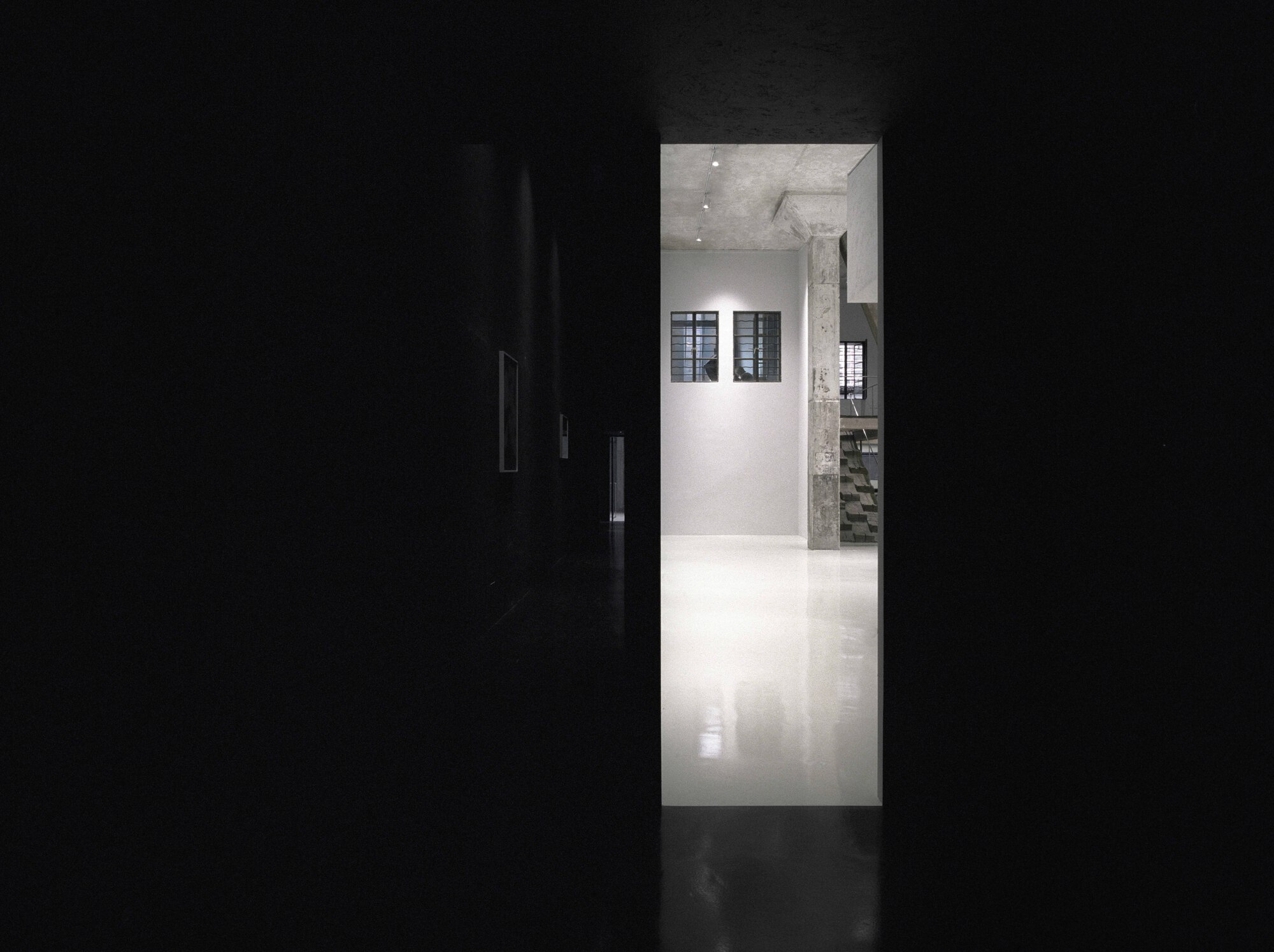 View through a dark corridor into a white-walled gallery space with concrete column and ceiling spotlights