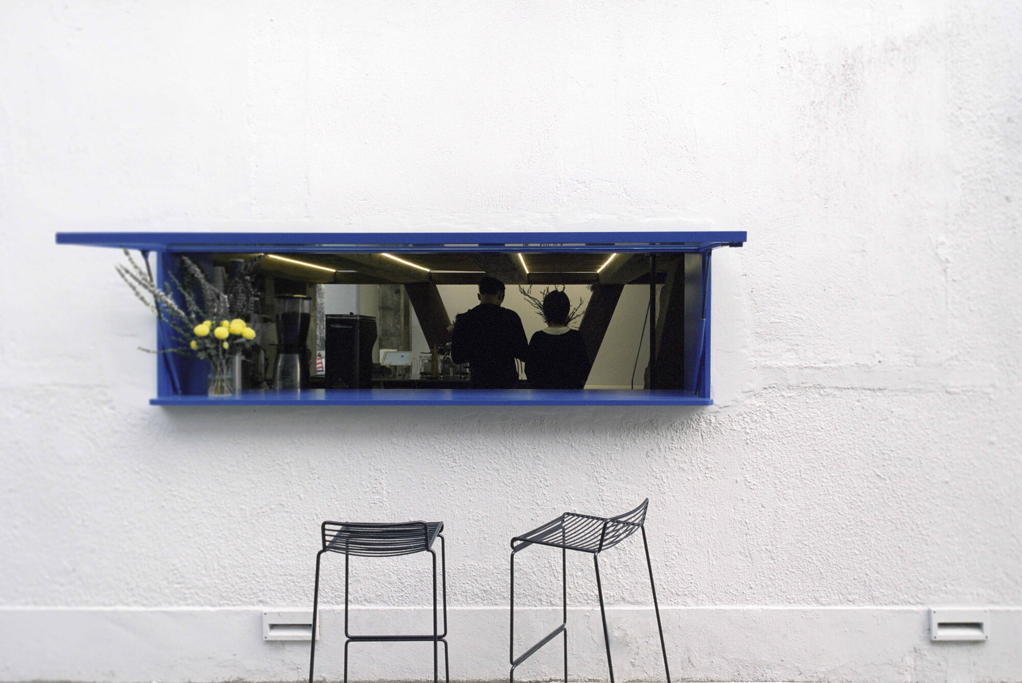 Blue-framed service window with mirrored interior set into white exterior wall above two wire stools