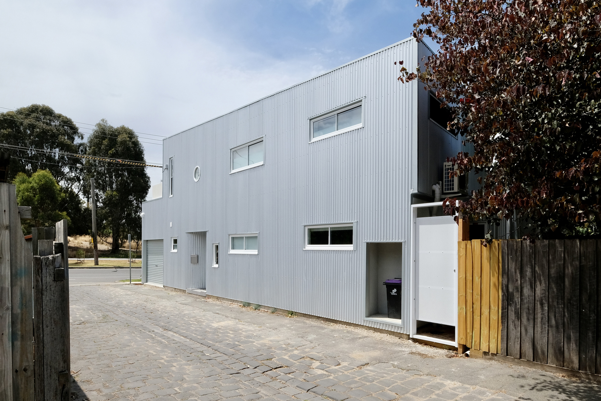 Side elevation with corrugated metal panels and a timber fence adjacent to the cobblestone lane
