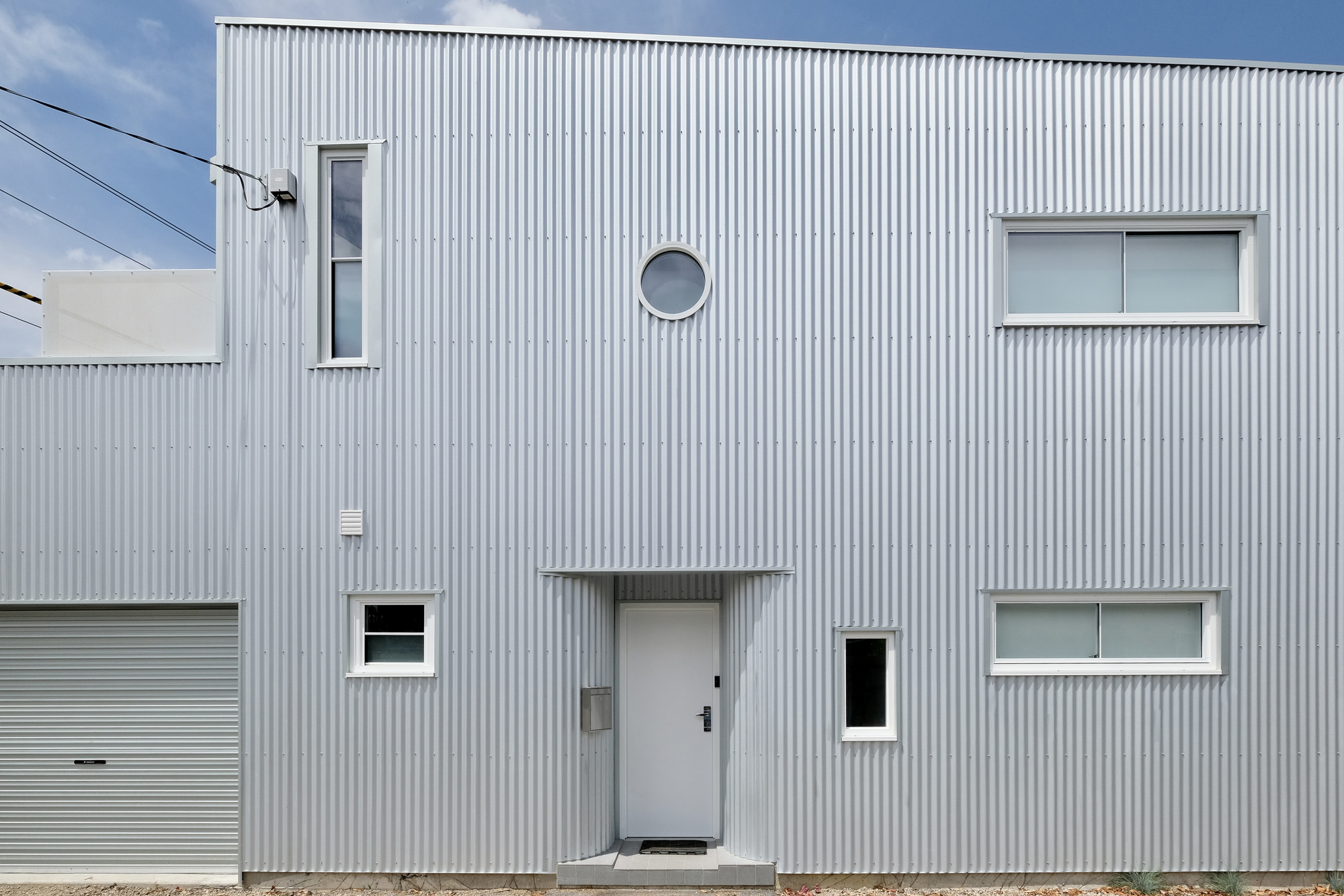 Front facade featuring a central door, circular porthole window, and scattered rectangular openings in corrugated cladding