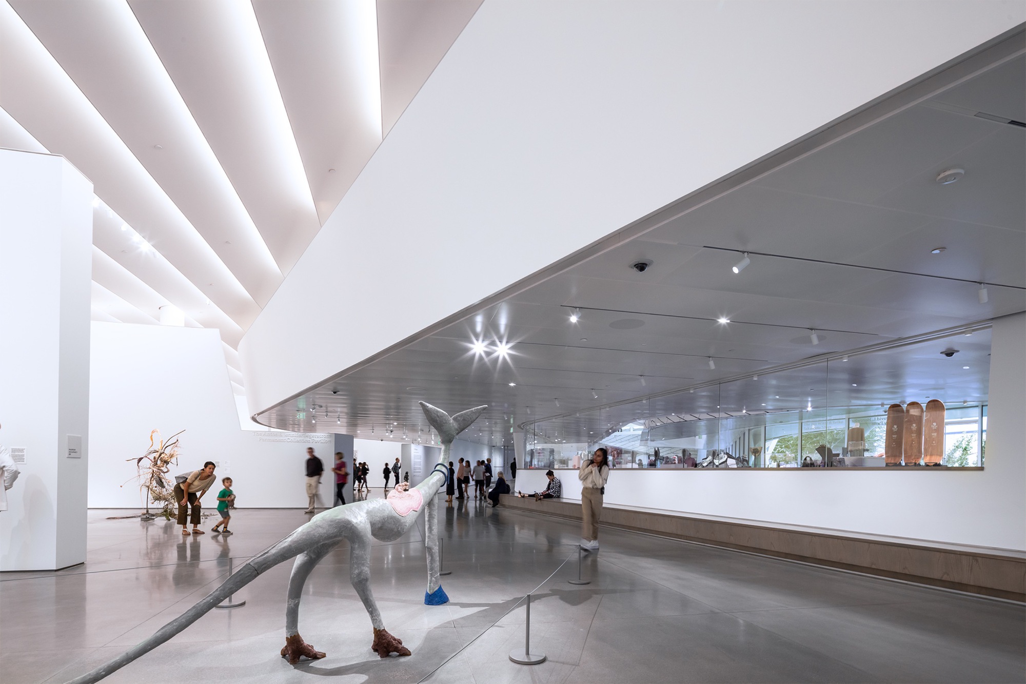 Gallery space with sculptural figures beneath a hovering metallic ceiling plane and skylights