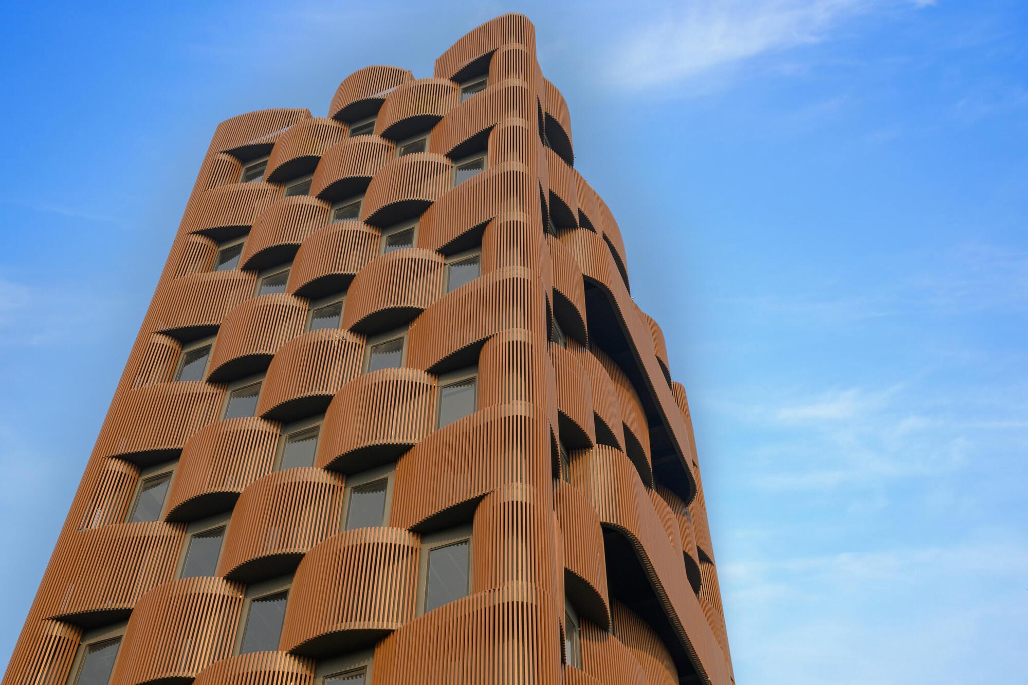 Tower facade clad in vertical terracotta fins with undulating corner geometry against wispy clouds
