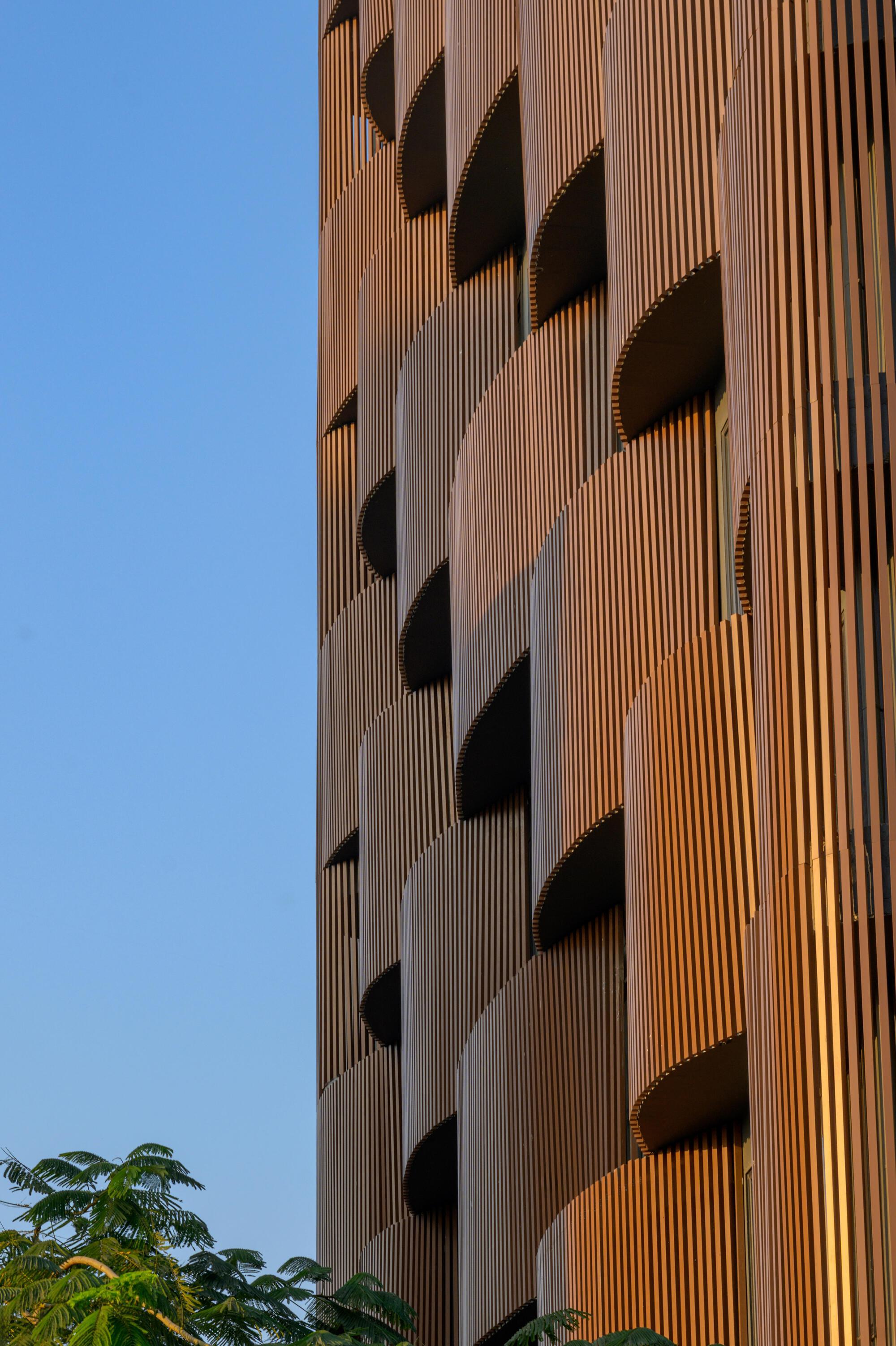 Detail of vertical terracotta fins wrapping the facade edge with tree foliage in foreground