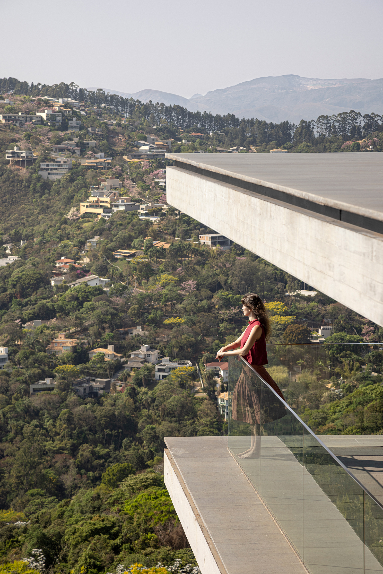 Cantilevered concrete terrace with glass balustrade overlooking a forested hillside dotted with houses