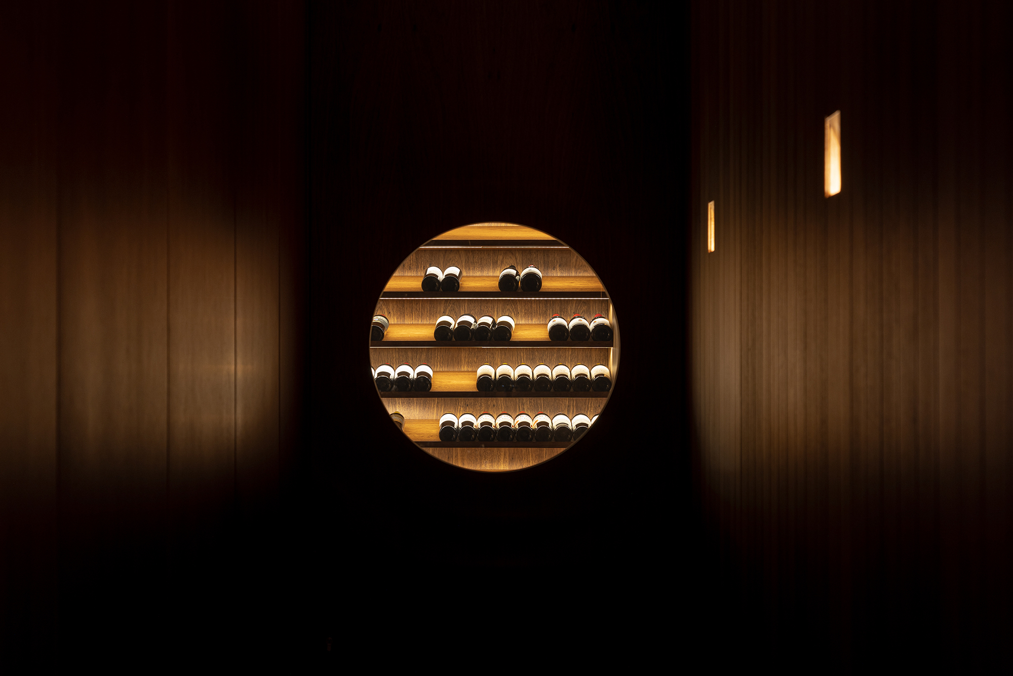 Circular aperture in a dark timber wall framing illuminated wine storage shelves