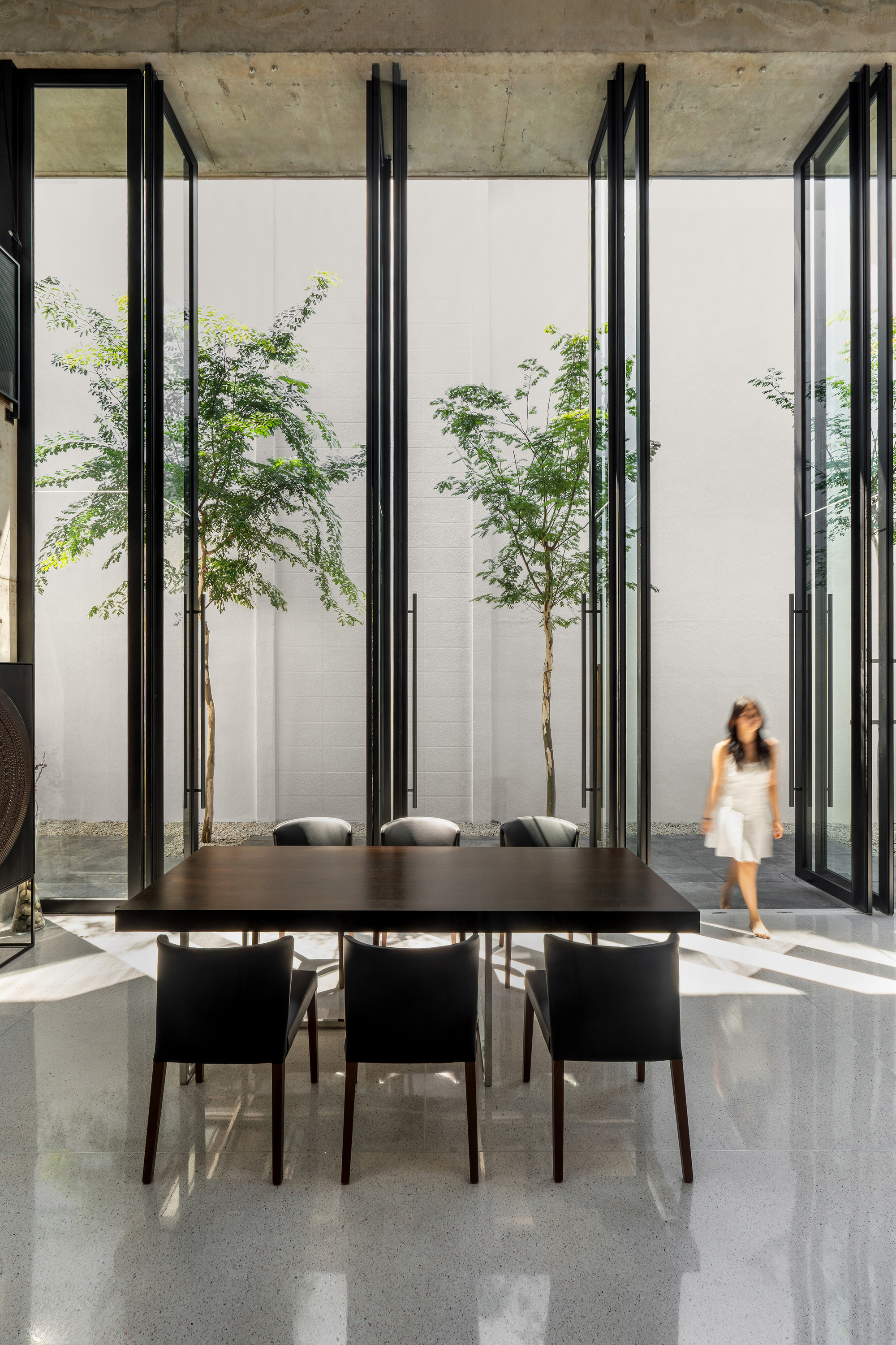 Black dining table beneath exposed concrete ceiling with tall glass walls framing courtyard trees and figure walking