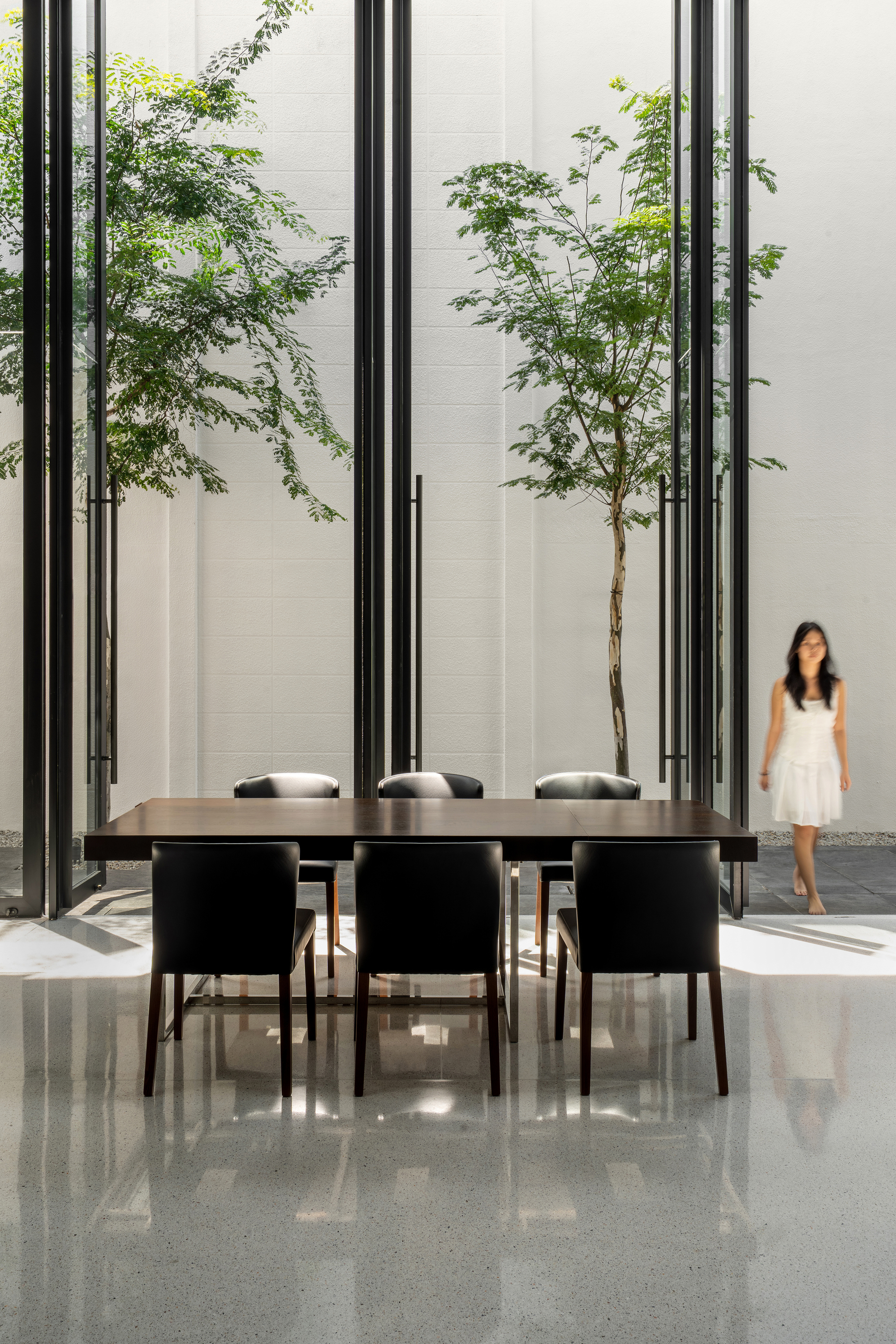 Dining table surrounded by steel frame screens with young trees and a figure in the courtyard