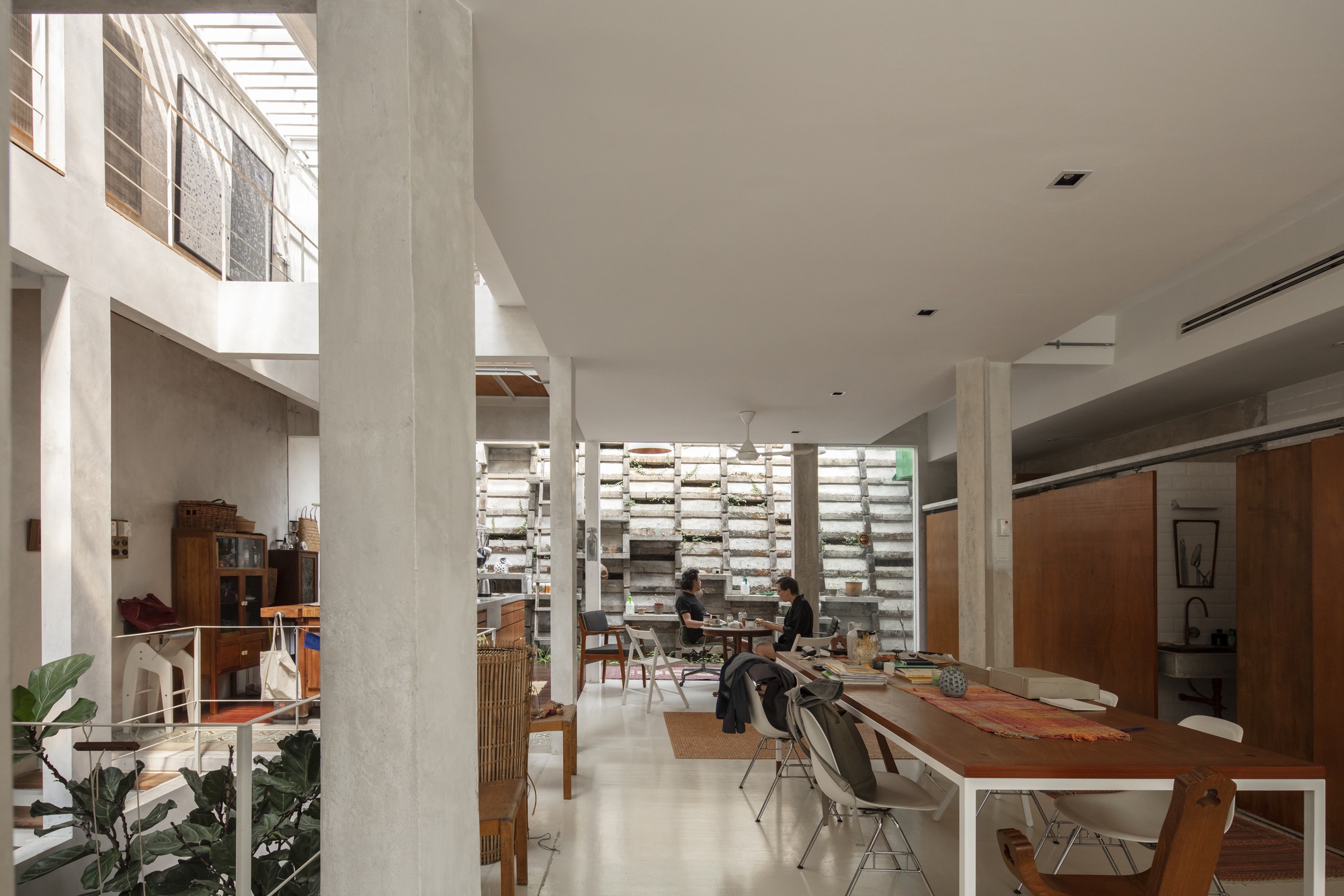 Double-height interior with concrete columns, timber dining table, and horizontal louvered screen wall with planted shelves