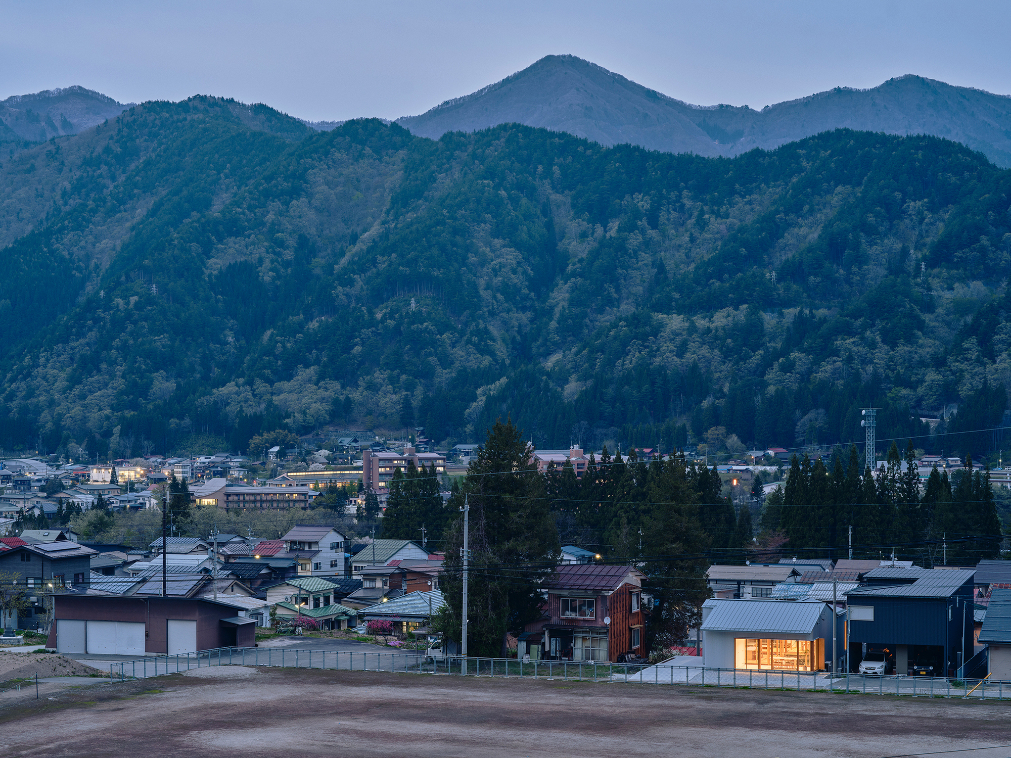 Illuminated single-story house with timber slatted facade at dusk in a village below forested mountains