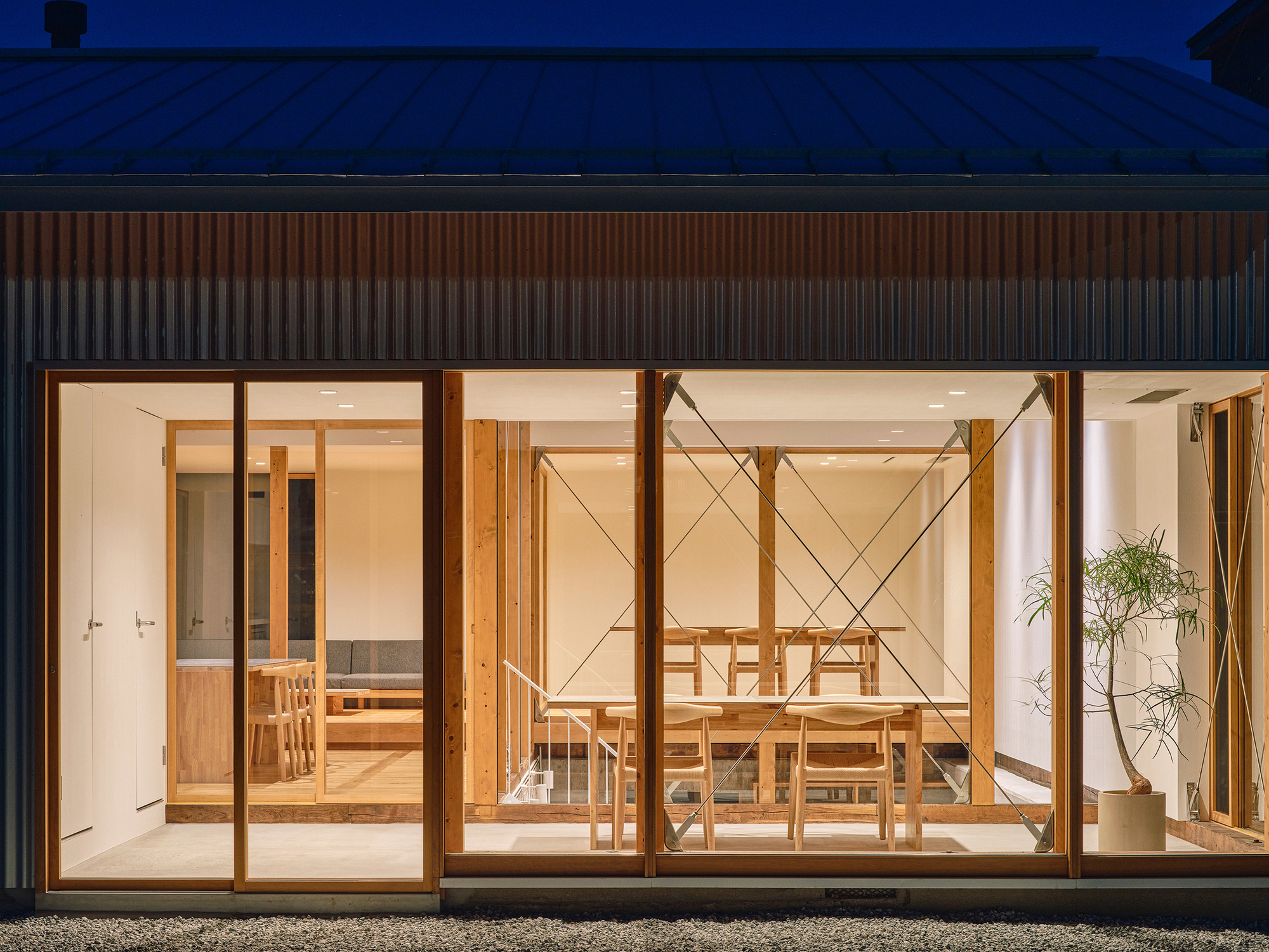 Glass wall revealing dining room with timber table and chairs beneath a corrugated metal roof at twilight
