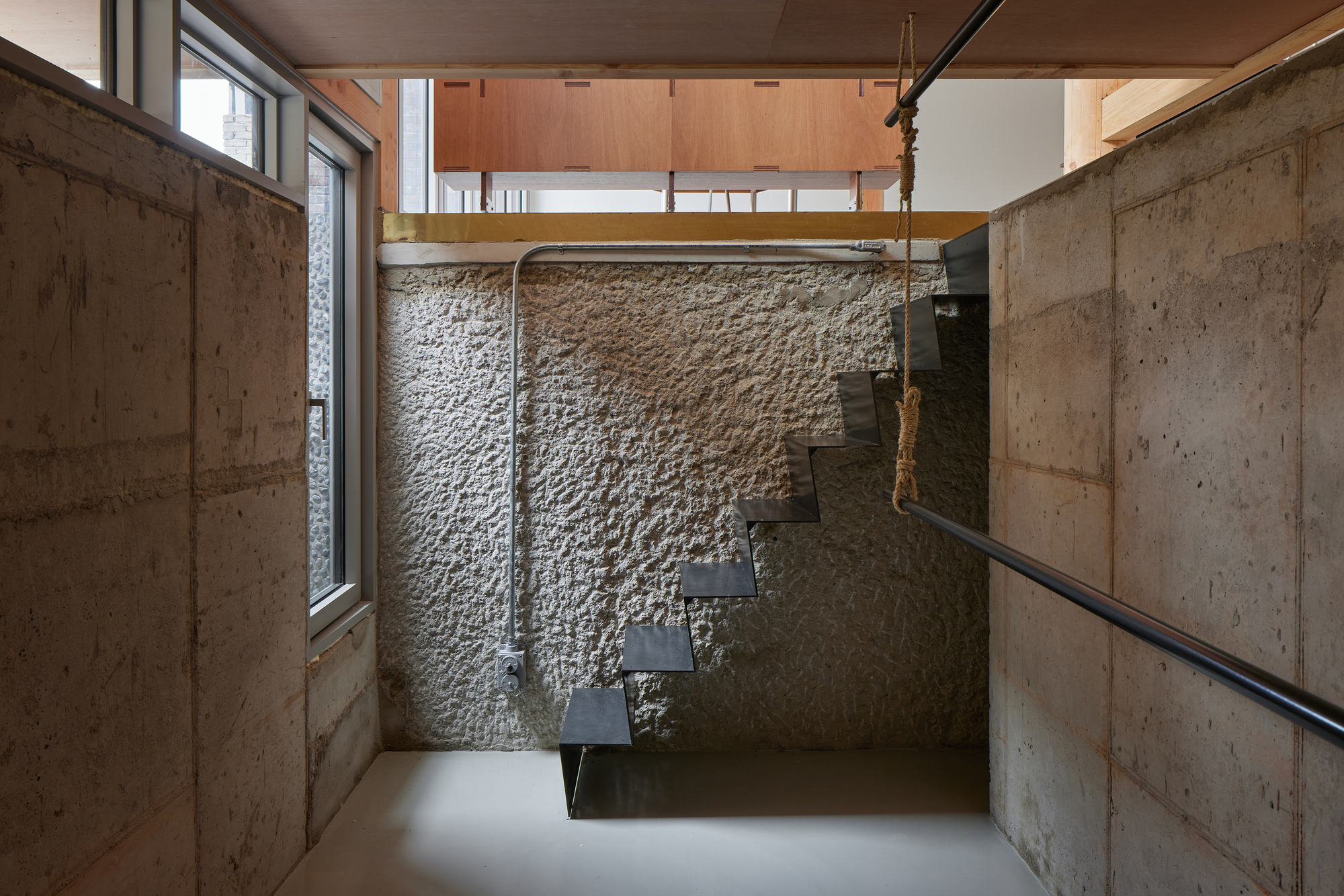 Floating steel staircase ascending against a textured stone wall flanked by concrete panels and rope handrails