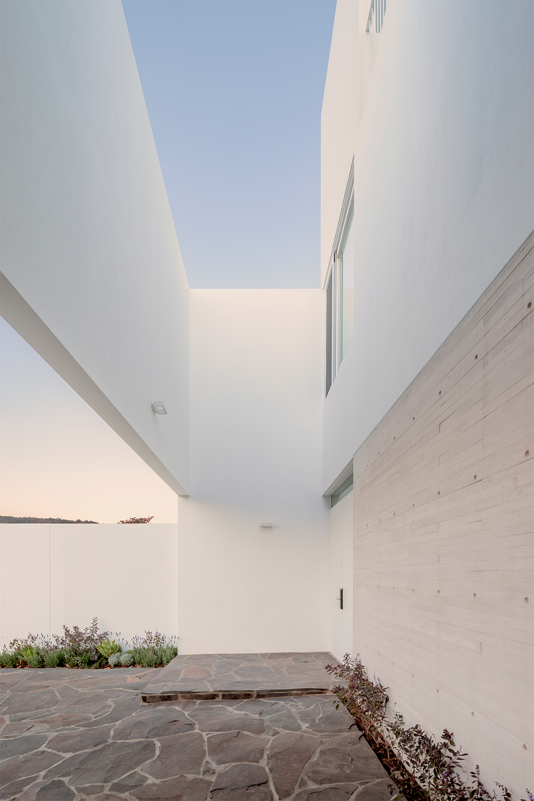Sloped passage with board-formed concrete wall and narrow skylight overhead at twilight
