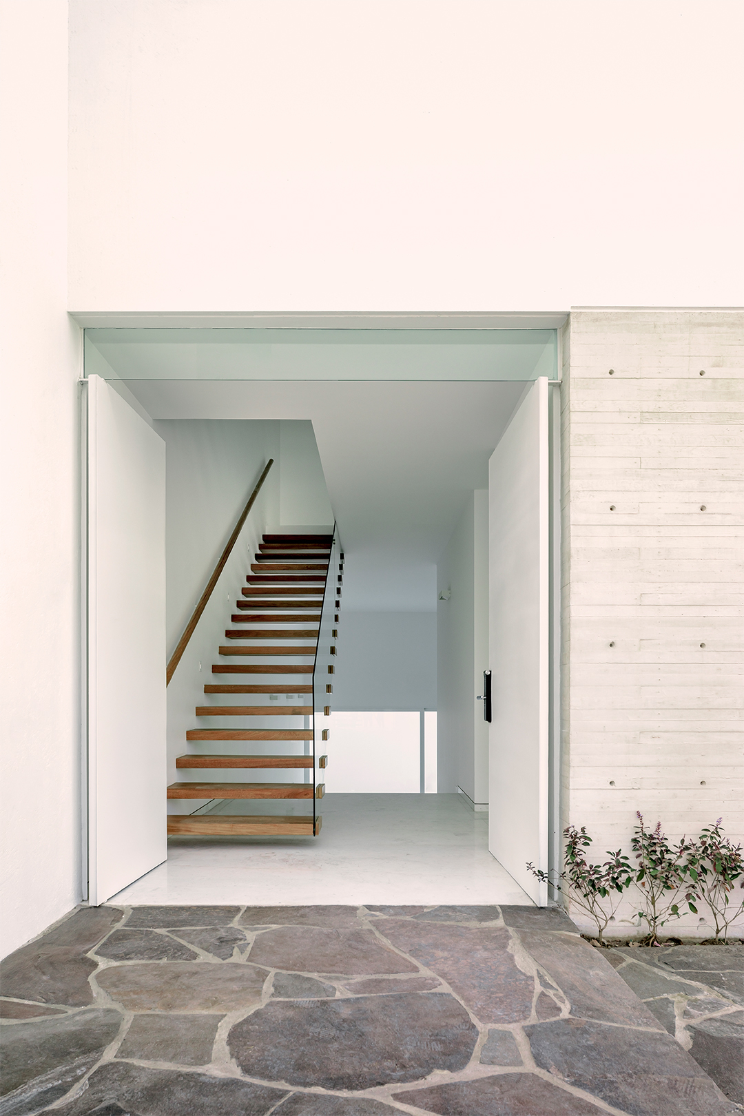 Entry threshold showing timber-tread staircase through open doorway beside board-formed concrete wall and stone paving