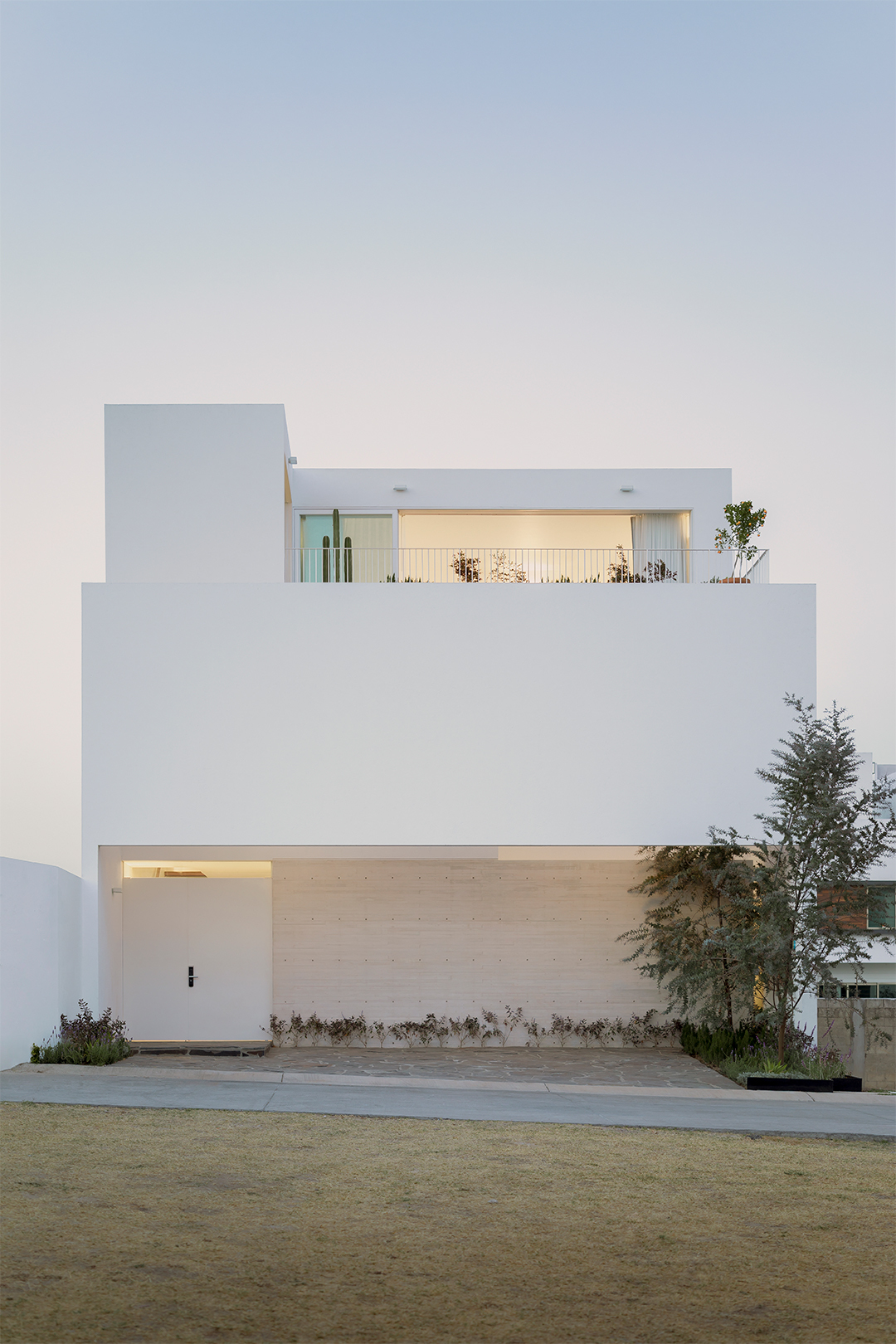 White-rendered facade with recessed garage and upper terrace flanked by young trees at dusk