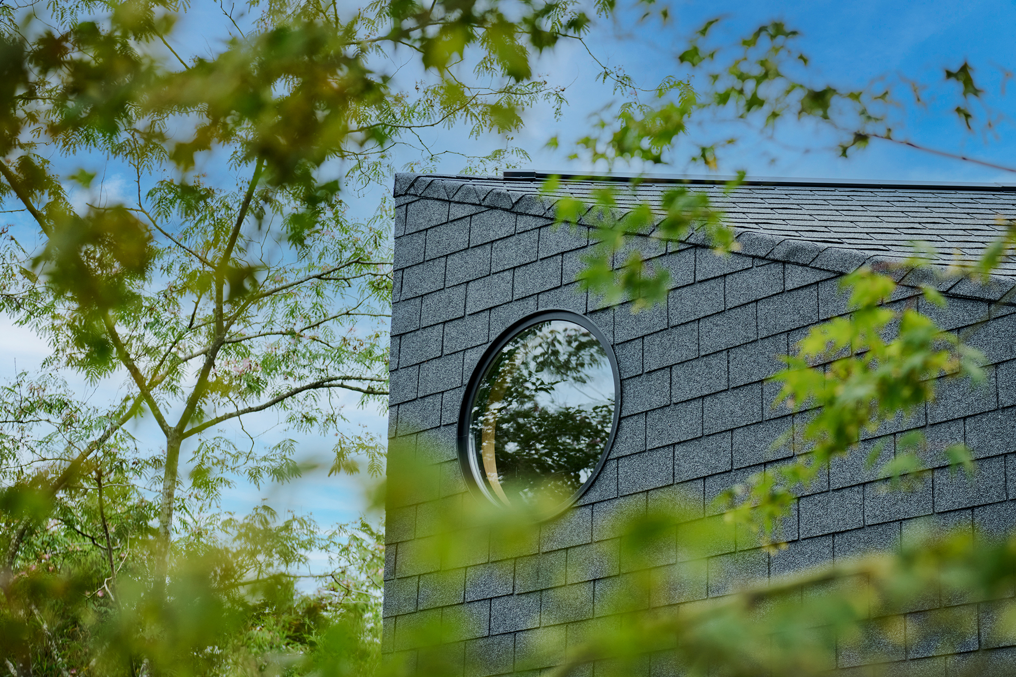 Circular window in grey shingled wall framed by spring leaves and blue sky
