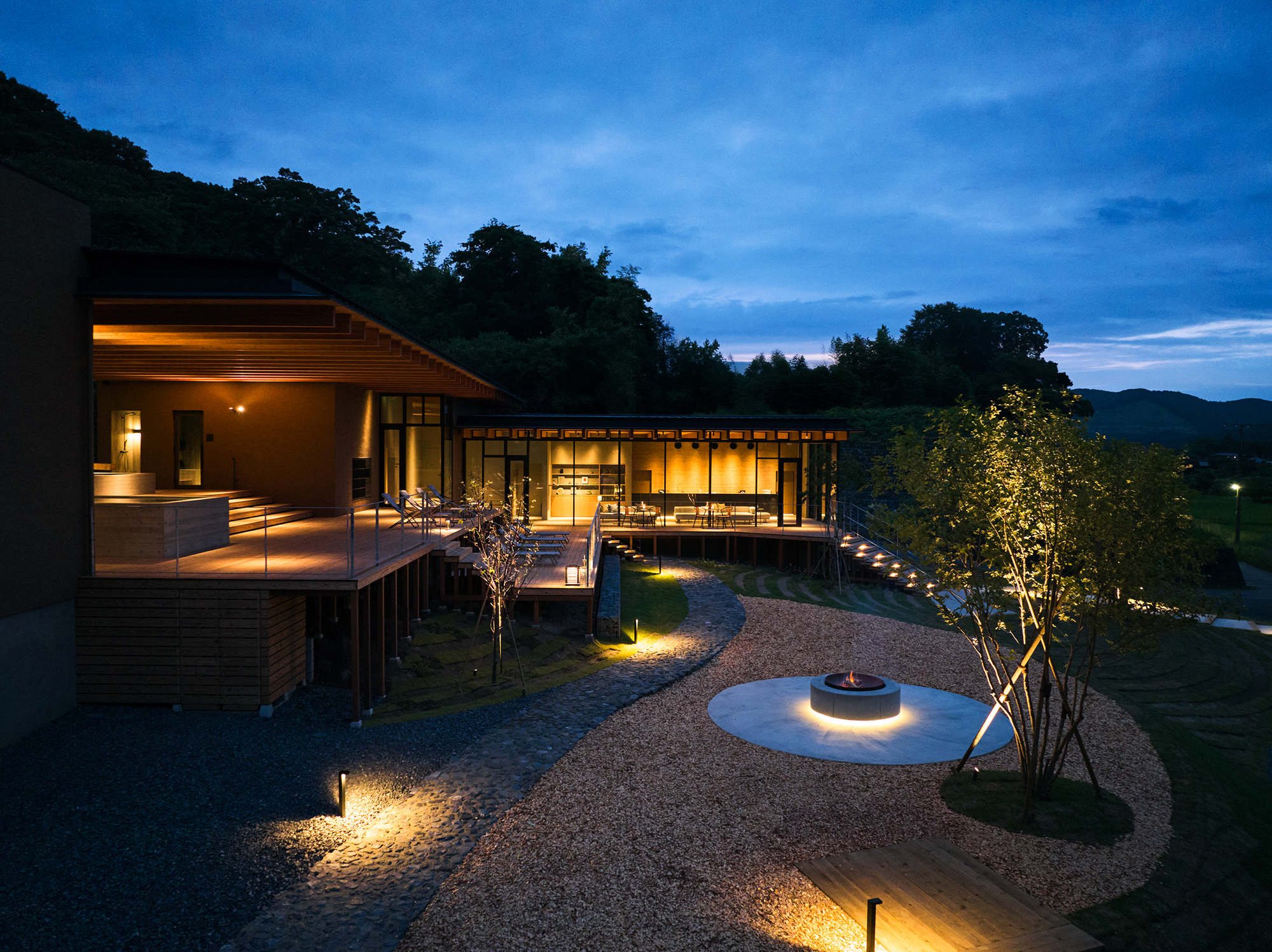 Glazed pavilion with timber deck and uplighting beneath a blue evening sky