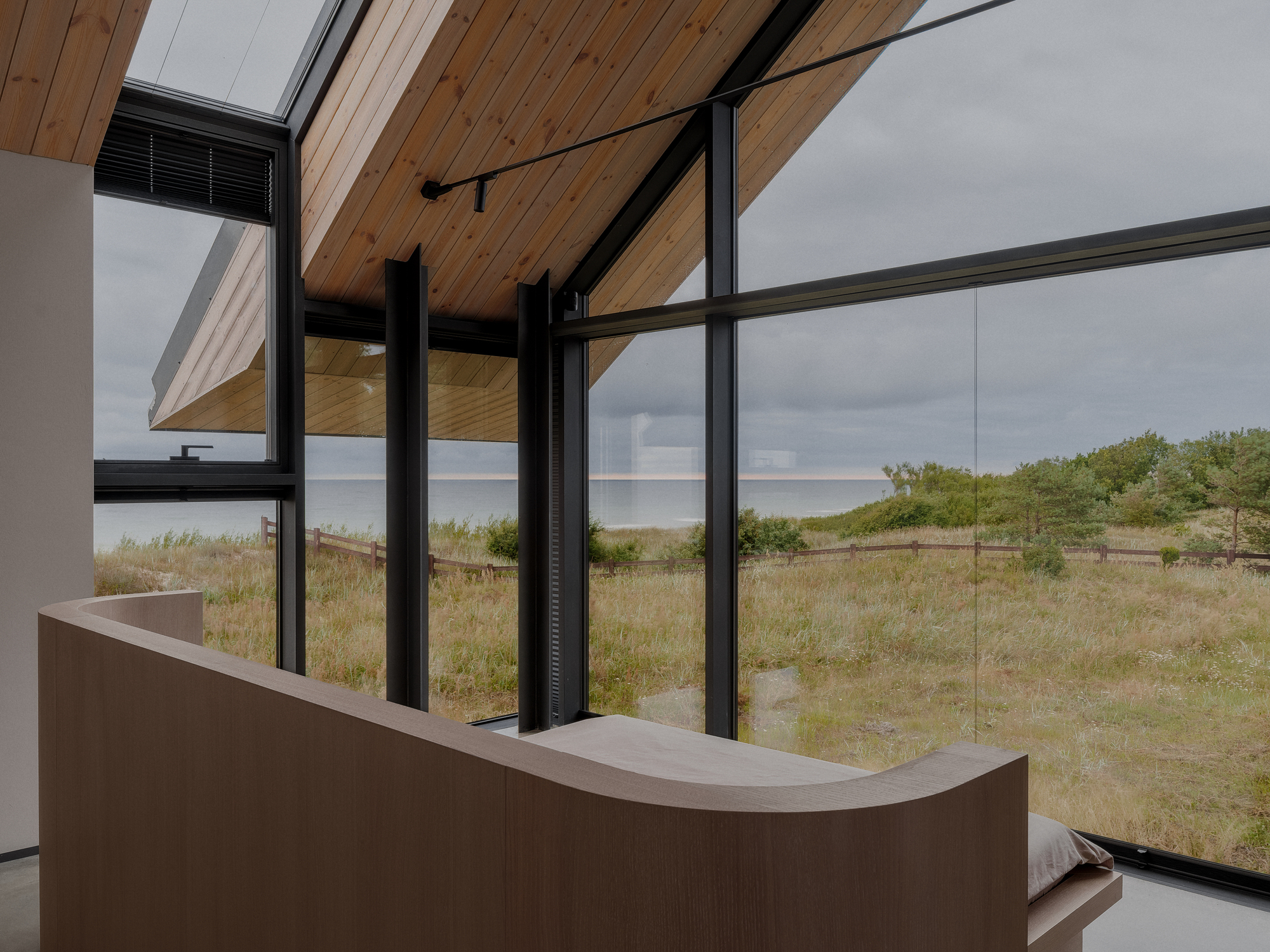 Interior corner with curved timber joinery and floor-to-ceiling black-framed glazing overlooking grassy dunes