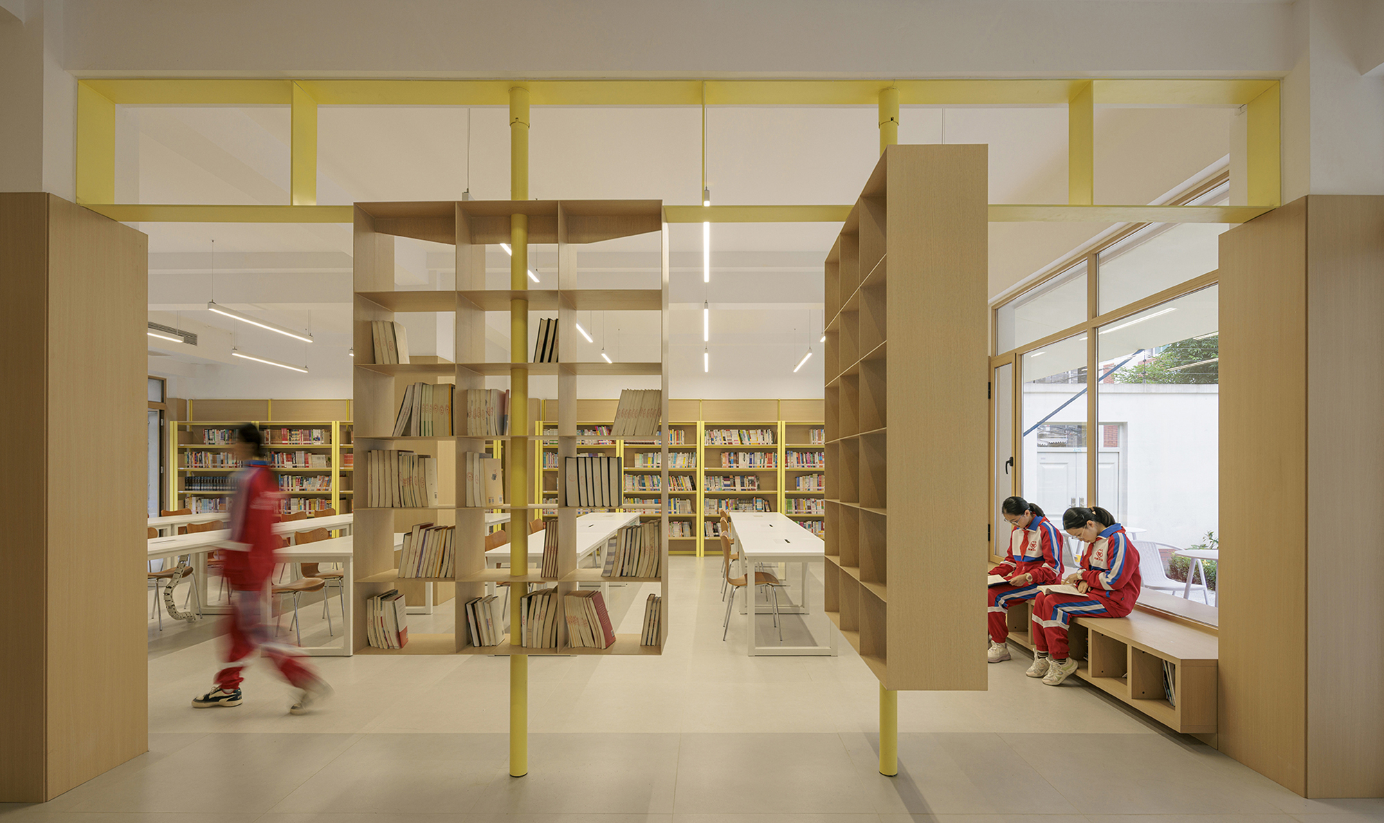 Library interior with a student walking past timber shelving and yellow steel framework near window seating