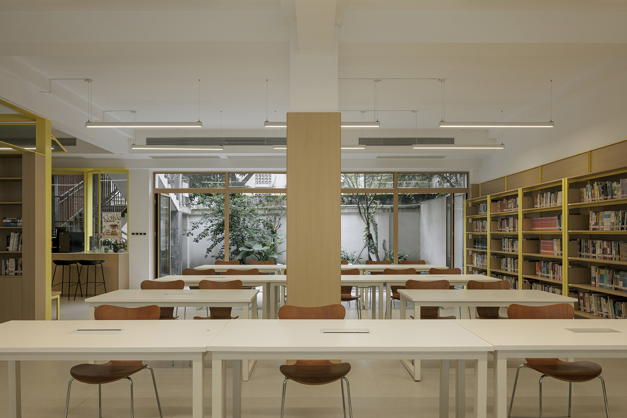 Reading room with white tables and timber stools facing courtyard windows framed by planters and trees