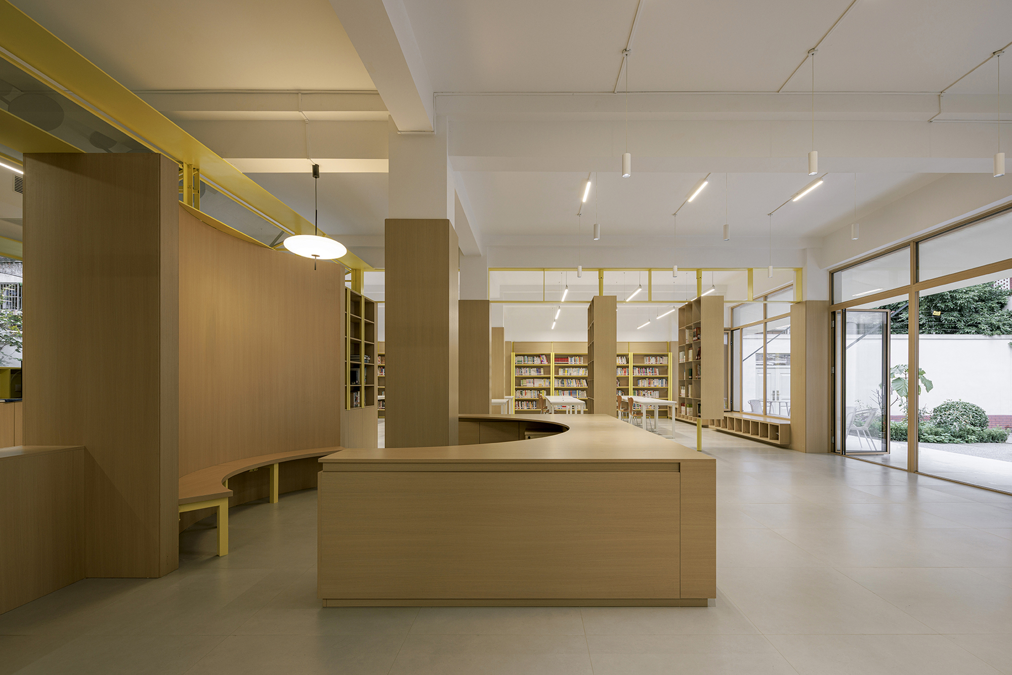 Central timber service counter beneath yellow ceiling beams with glazed doors opening to a courtyard beyond