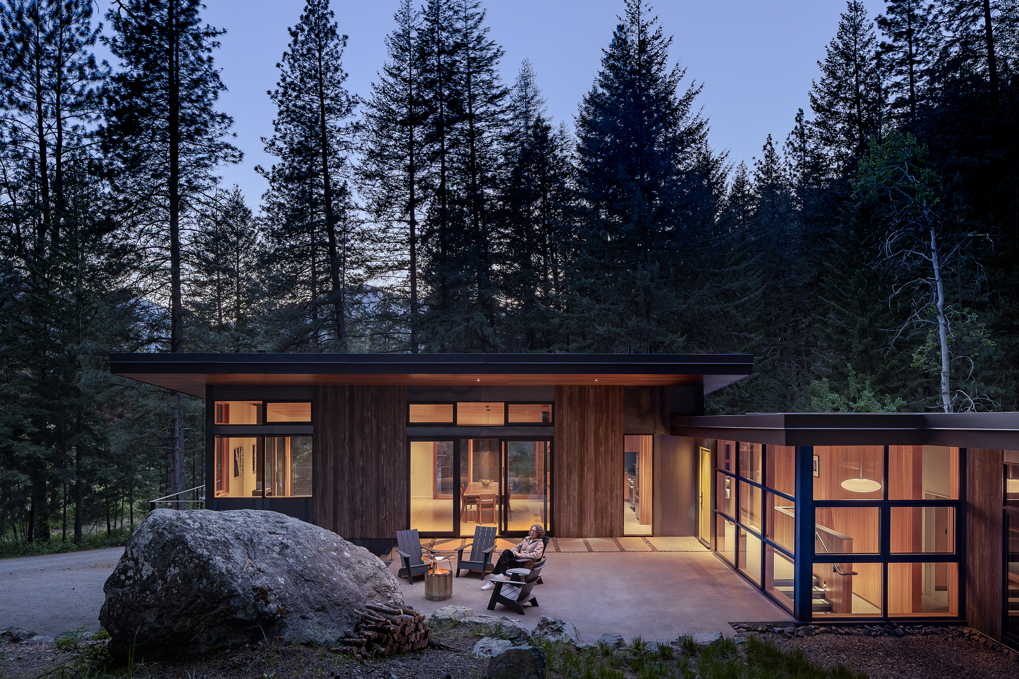 Timber-clad volumes with flat roofs and glazed walls glowing at dusk among tall conifers