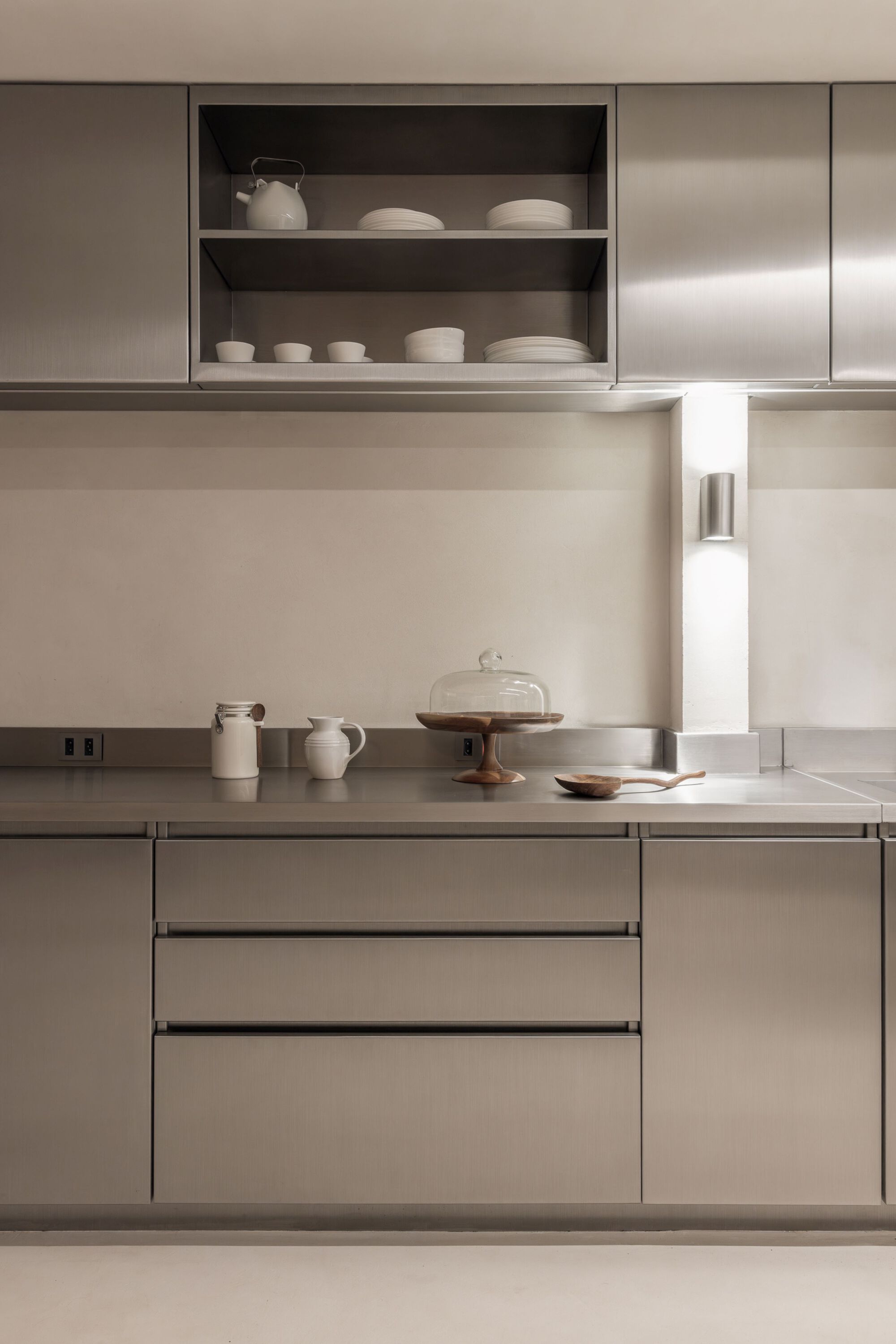 Stainless steel countertop with open shelving displaying ceramic vessels and a recessed task light
