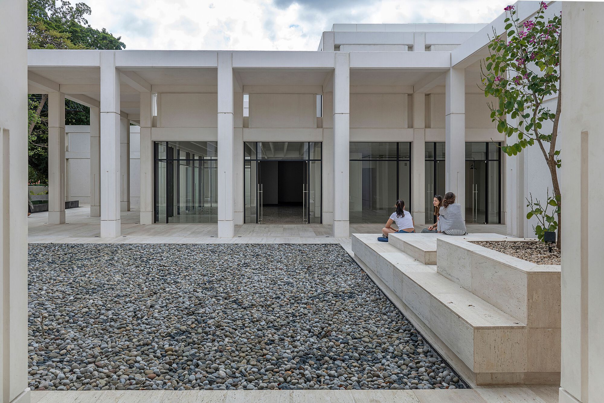 Courtyard with gravel bed and concrete planter bench where two visitors sit beneath a colonnade