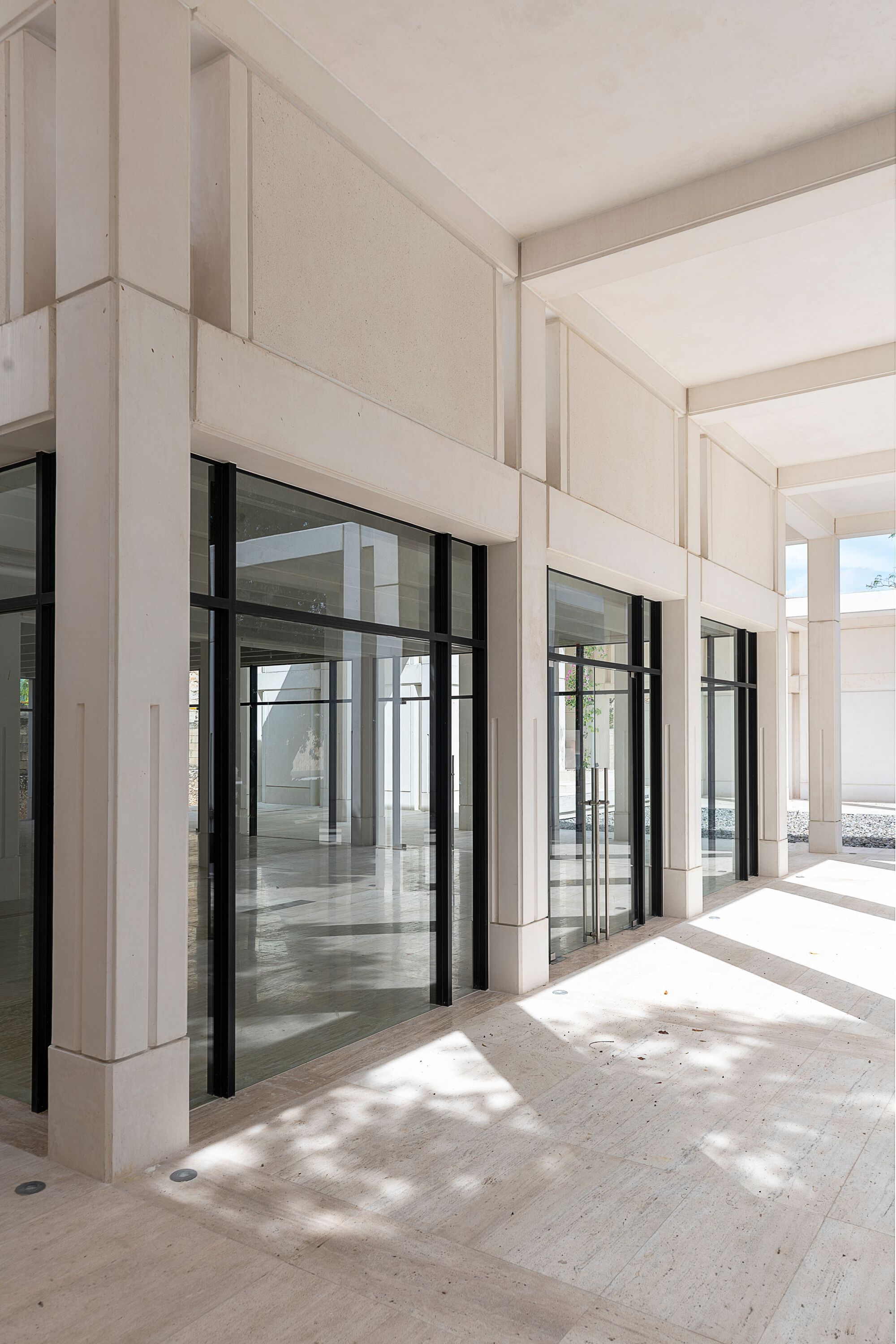 Covered portico with pale stone columns and black-framed glass doors casting dappled shadows on the floor