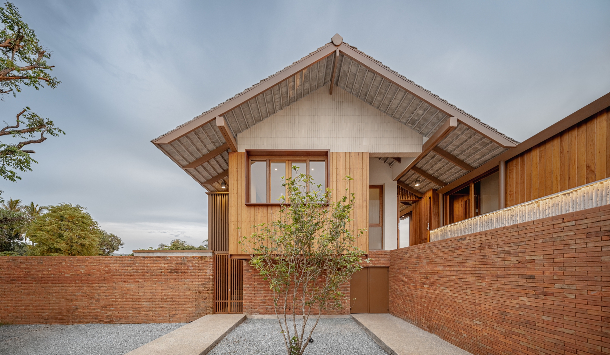Entry courtyard framed by brick walls with a young tree and timber-clad gabled volume under overcast skies