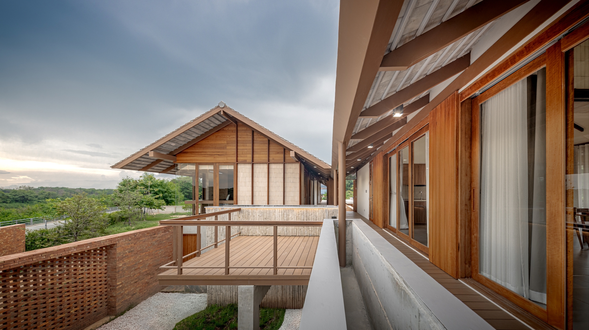 Elevated walkway with metal railings linking two gabled volumes overlooking green landscape at dusk