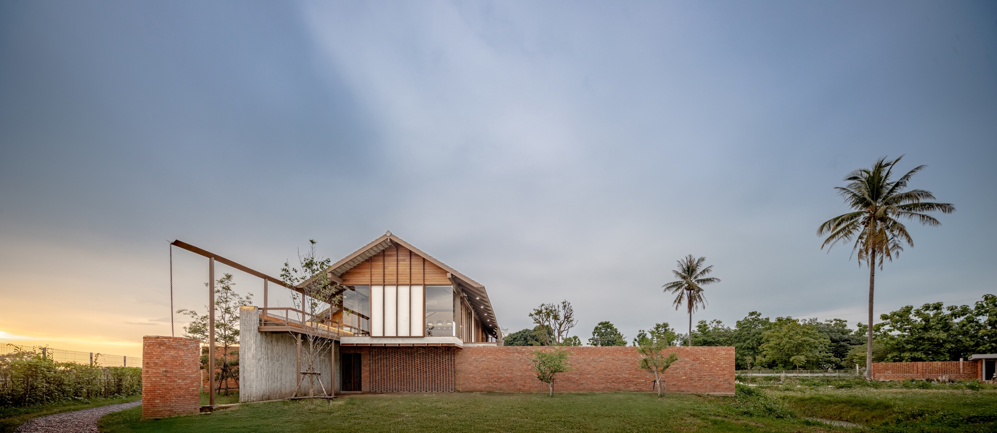 Gabled volume with timber cladding rising above brick perimeter wall at sunset with palm trees