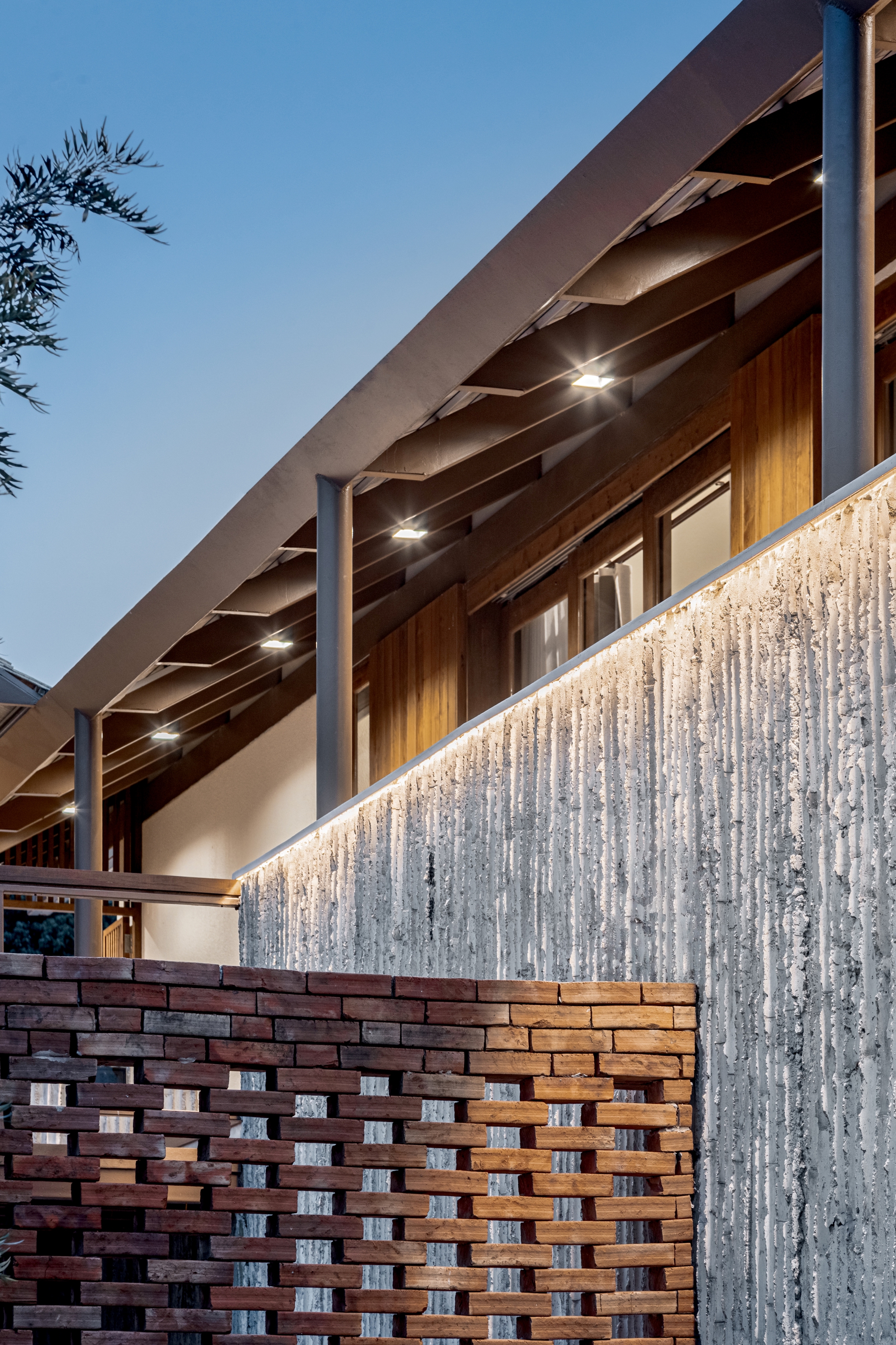 Sloping exposed timber roof over a woven brick screen wall and textured concrete at dusk