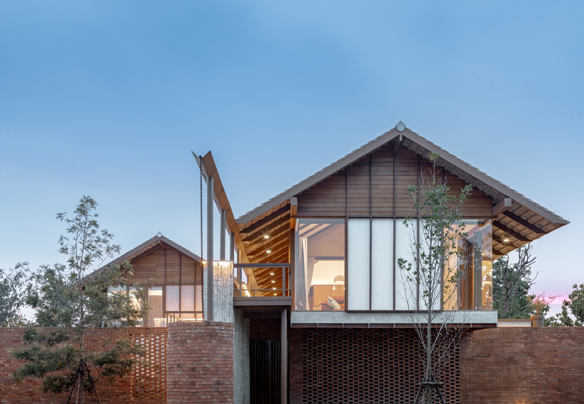 Twin gabled volumes with glazed facades and timber louvers rising above a perforated brick wall at dusk