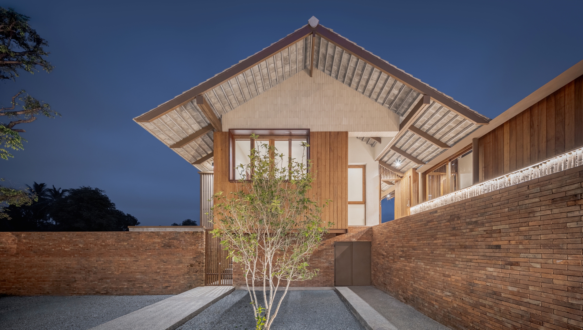 Timber gable volume with overhanging eaves rising above a brick courtyard wall at dusk