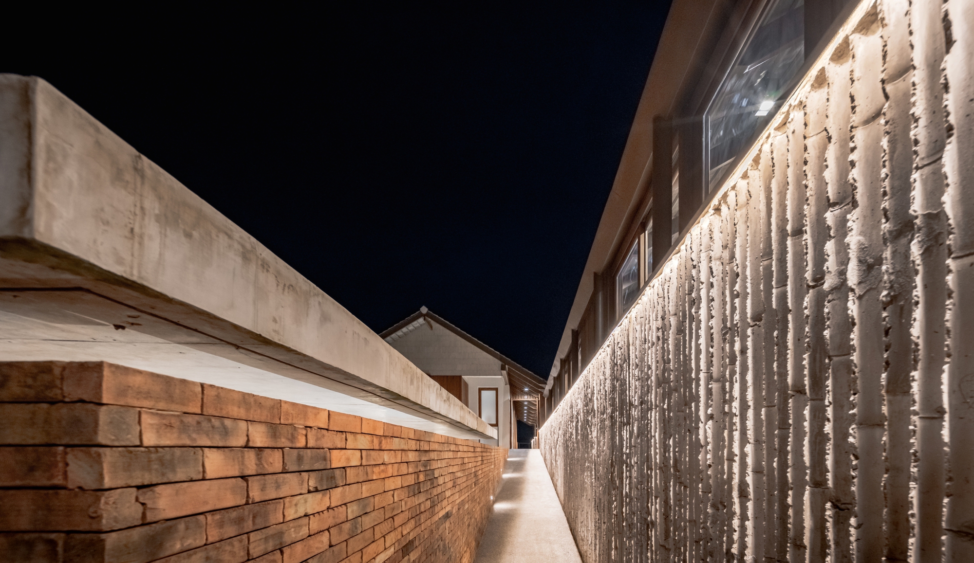 Narrow concrete walkway between brick wall and illuminated perforated screen facade at night