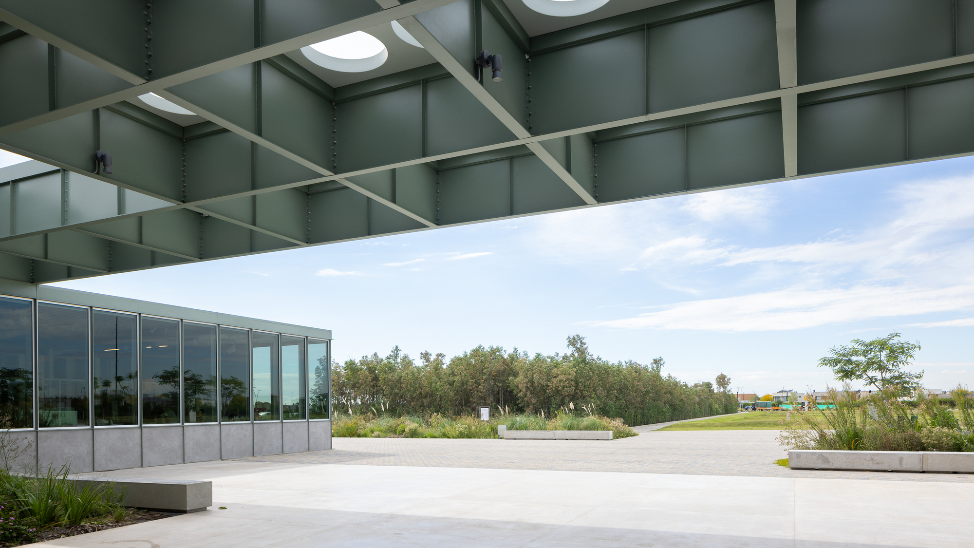 Covered terrace with green steel canopy and skylights overlooking a concrete plaza and planted landscape