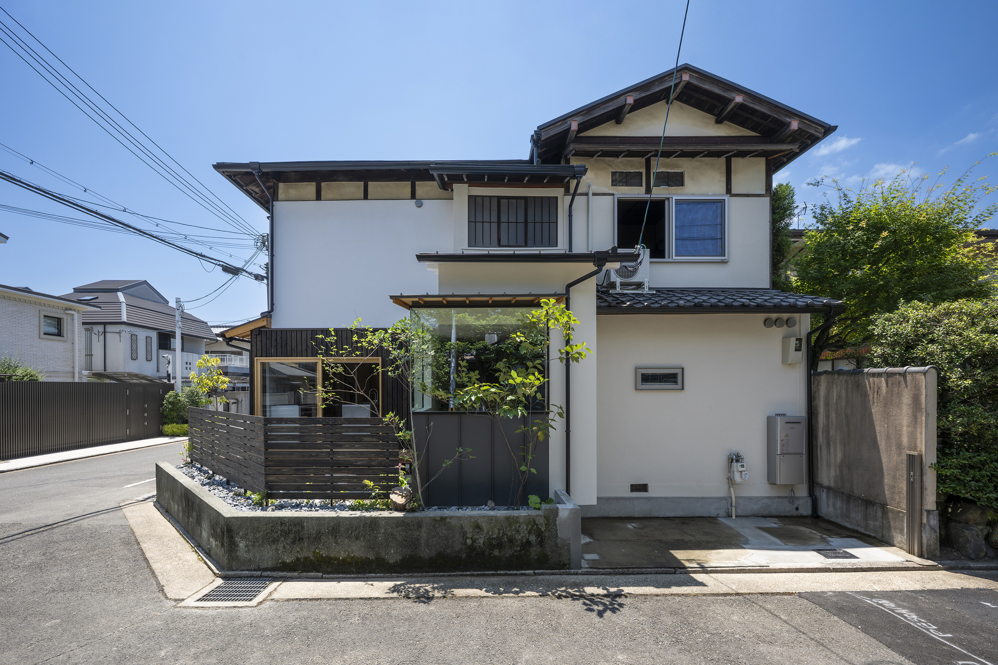 Street facade with white stucco walls, dark horizontal screens and small planted garden on a clear day