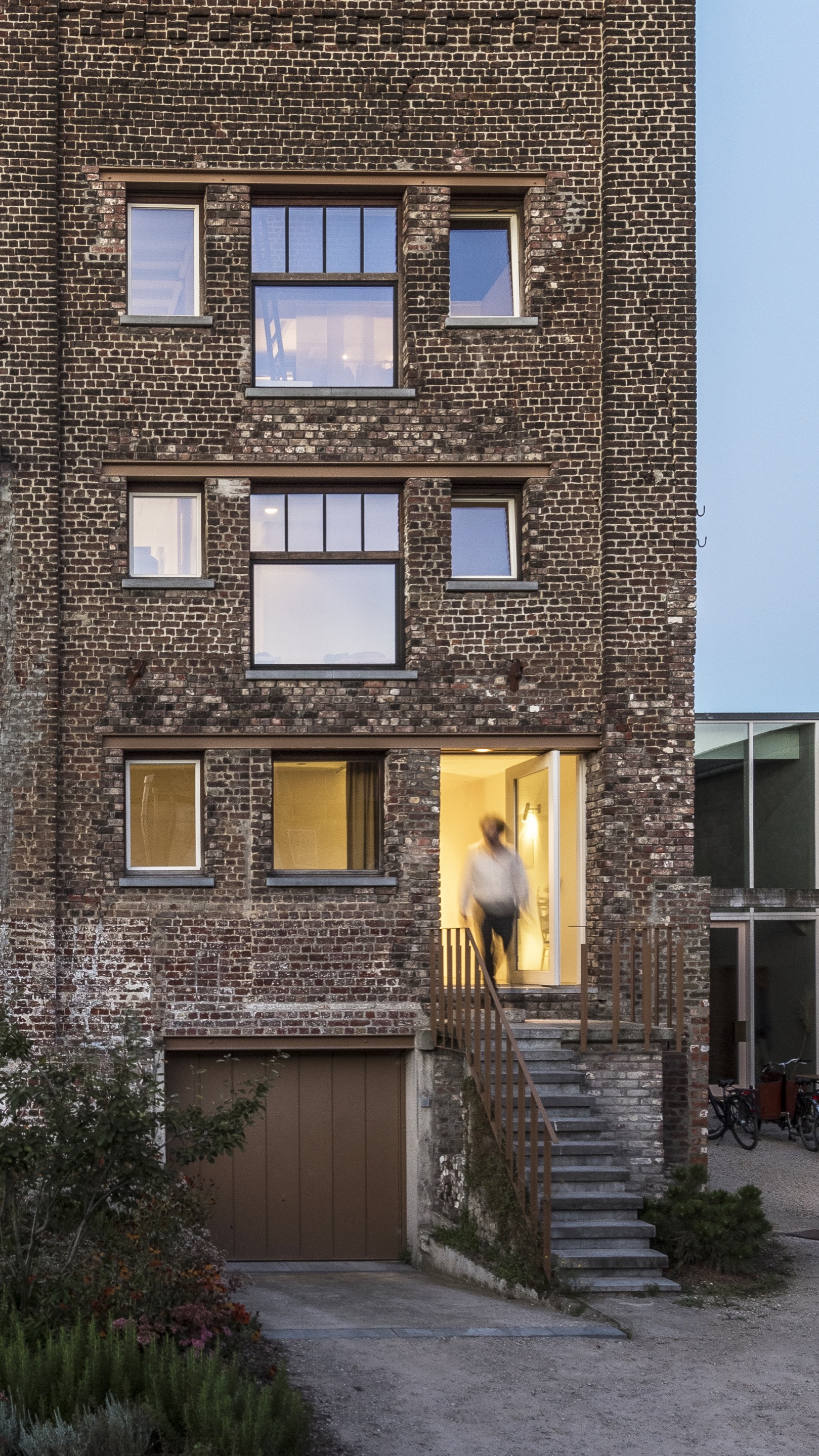 Facade at dusk with reclaimed brick wall and illuminated entry doorway at top of stone stair