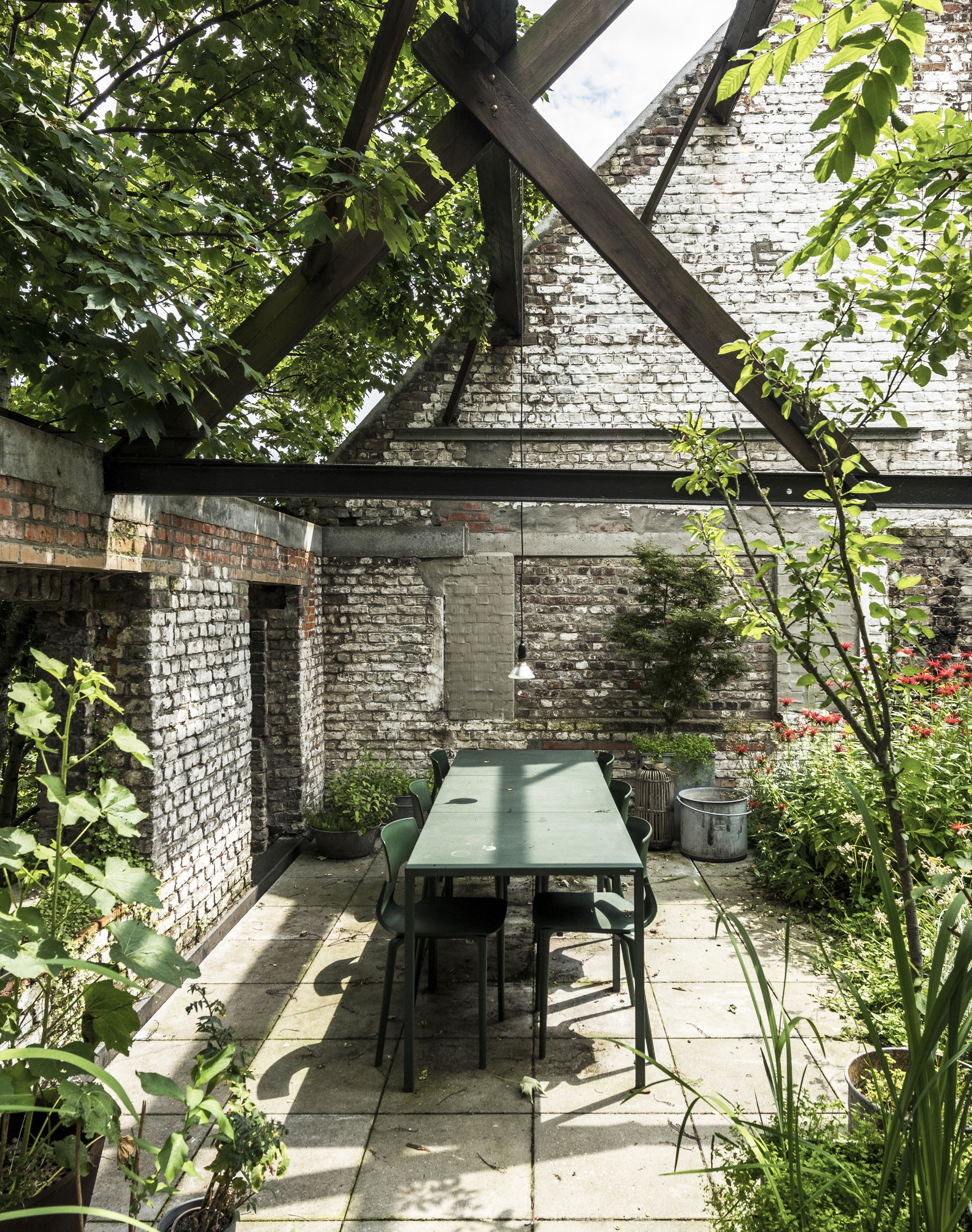 Outdoor dining table under timber roof frame with whitewashed brick gable wall and dappled tree shade