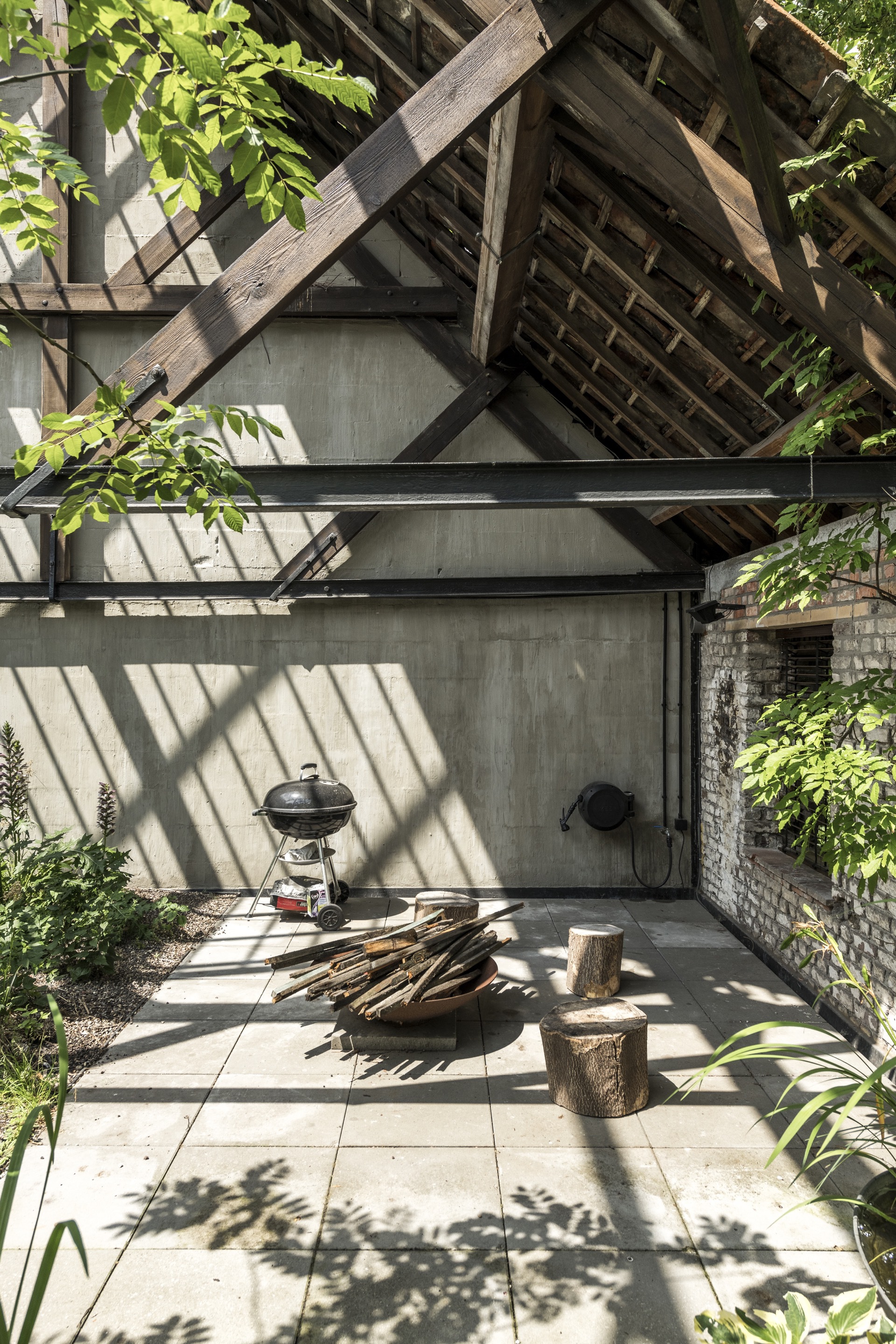 Paved courtyard with charcoal grill and firewood beneath angled timber structure casting diagonal shadows