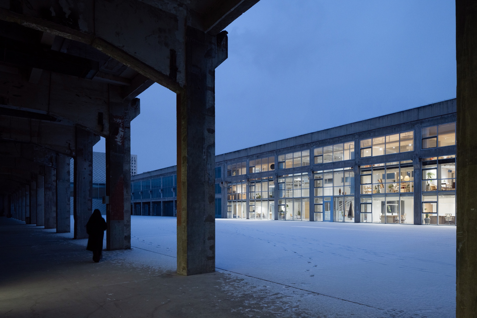 View through concrete columns toward illuminated glass facade across a snowy plaza at twilight