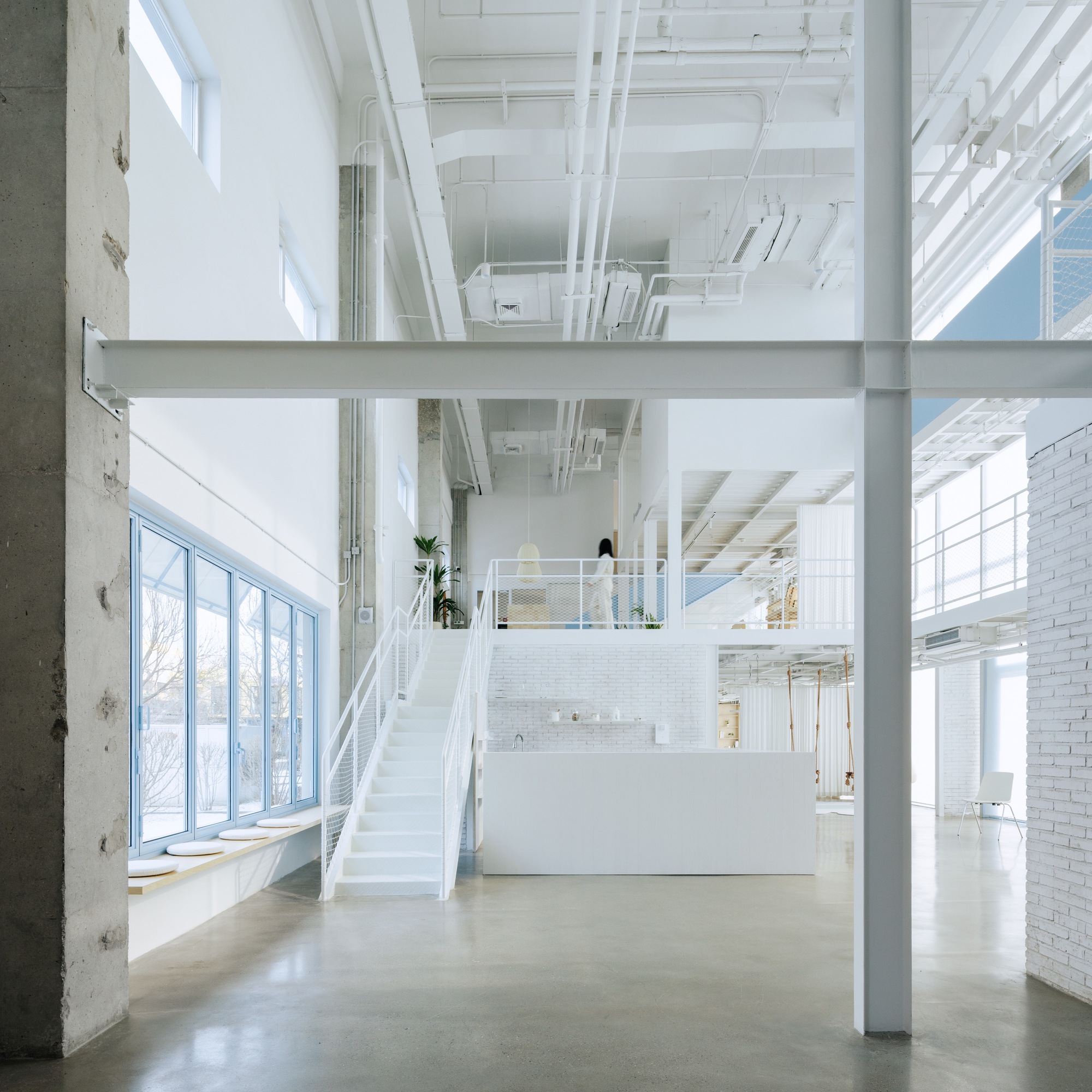 Double-height interior with white steel stair connecting mezzanine levels under exposed mechanical systems