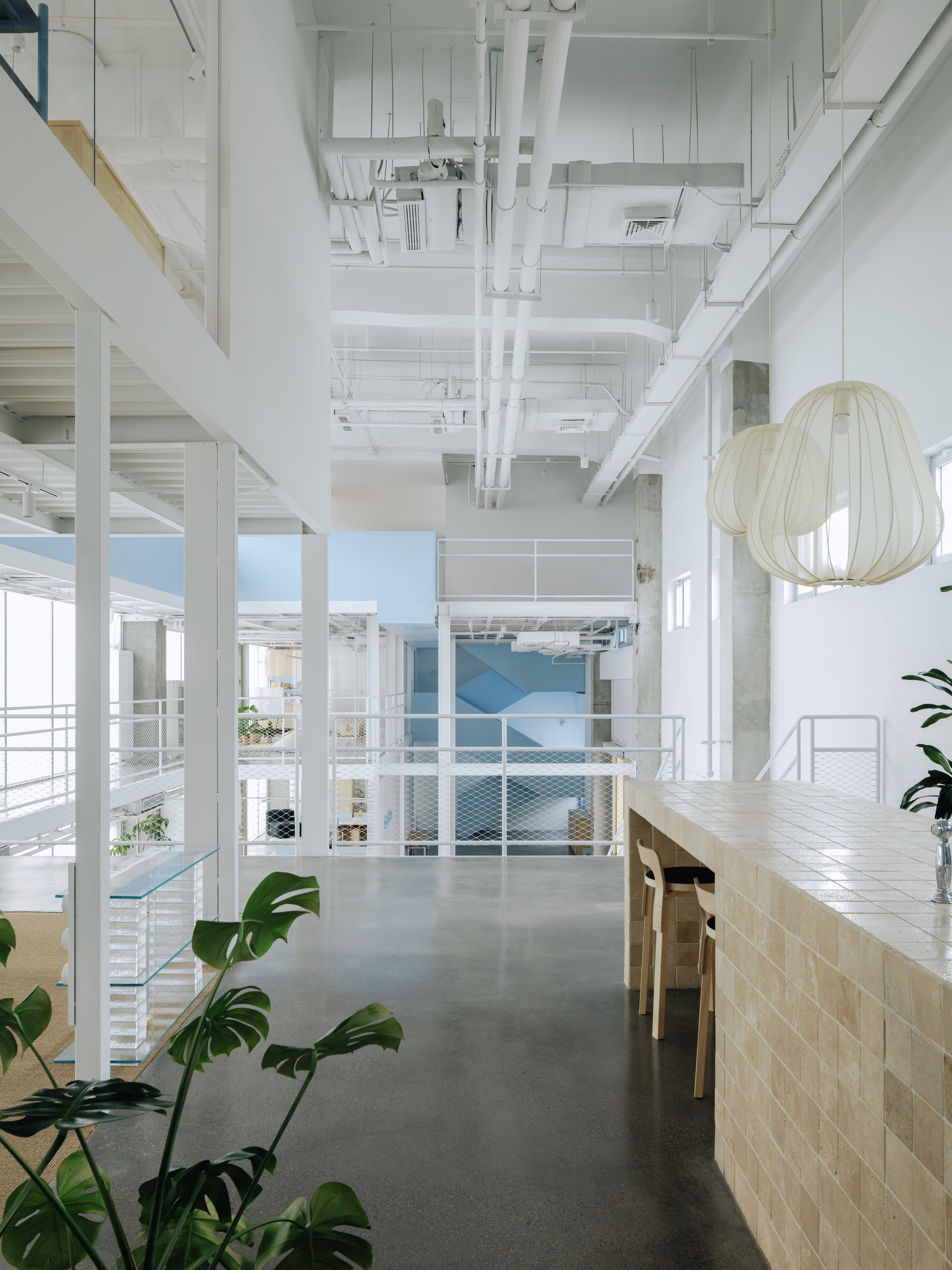 Double-height corridor with exposed white mechanical systems, metal railings, and potted plants in sunlight