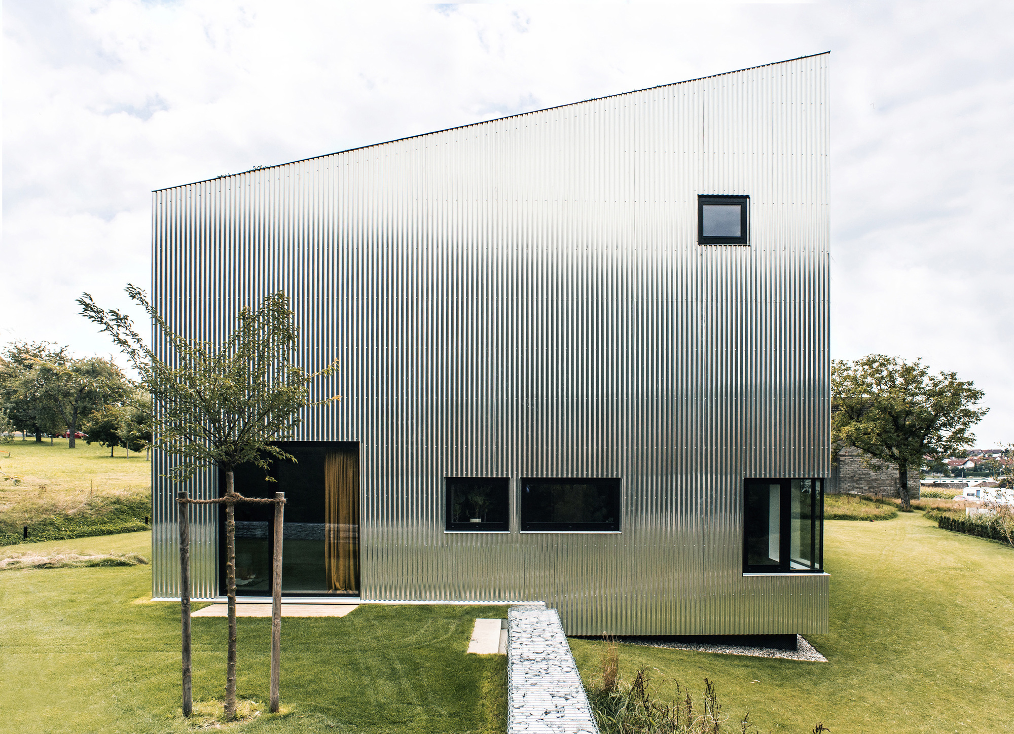 Corrugated metal box volume elevated on the sloping lawn with young trees planted nearby