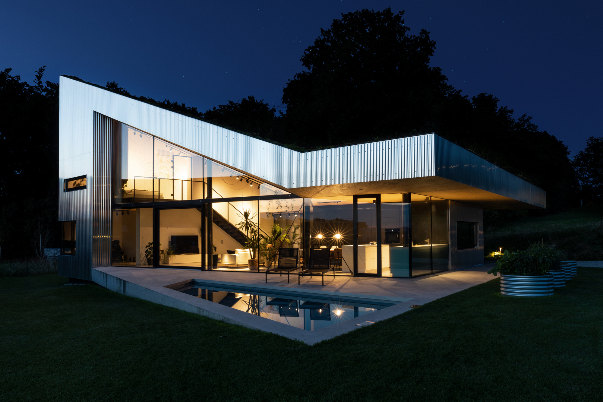 Night view of the corrugated metal roof extending over the glazed living area and terrace with pool