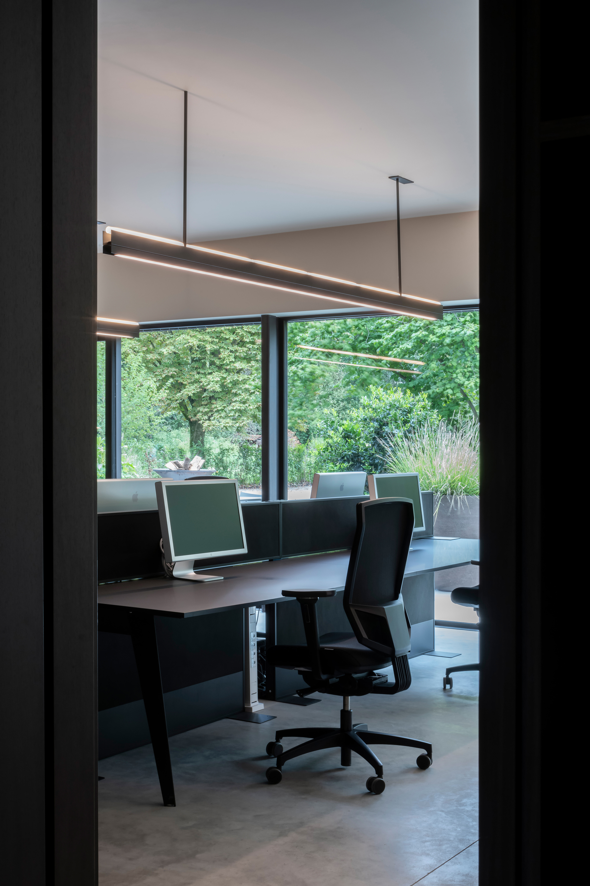 Office desk framed by doorway with suspended linear lighting and garden view beyond