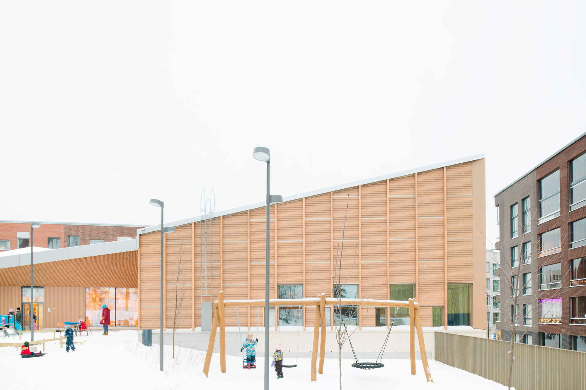 Timber facade with ground-level glazing overlooking a snow-covered playground with children and swings