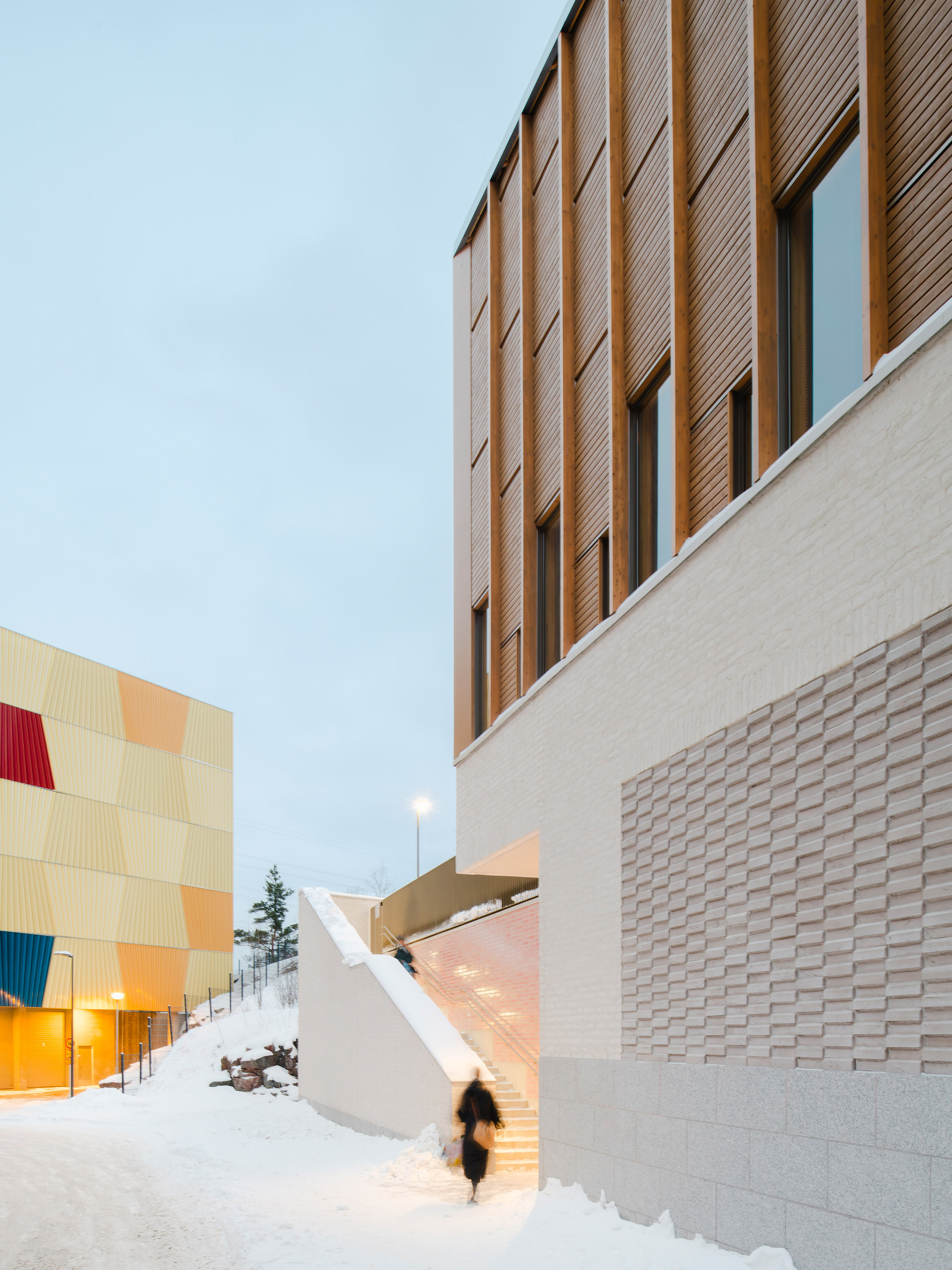 Exterior passage between textured brick base and vertical wood cladding during winter twilight