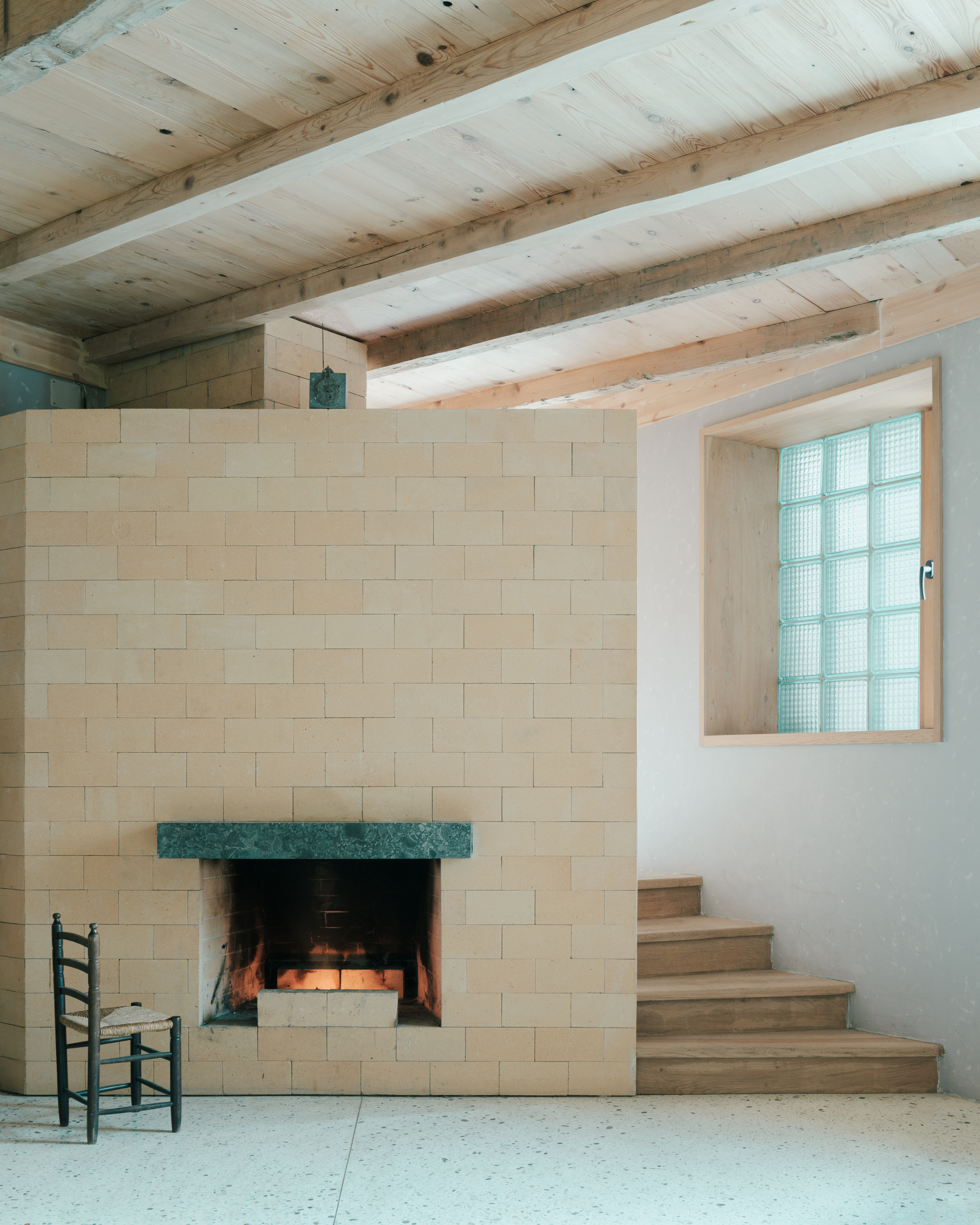 Pale stone fireplace with green marble mantel beneath timber ceiling joists and glass block window
